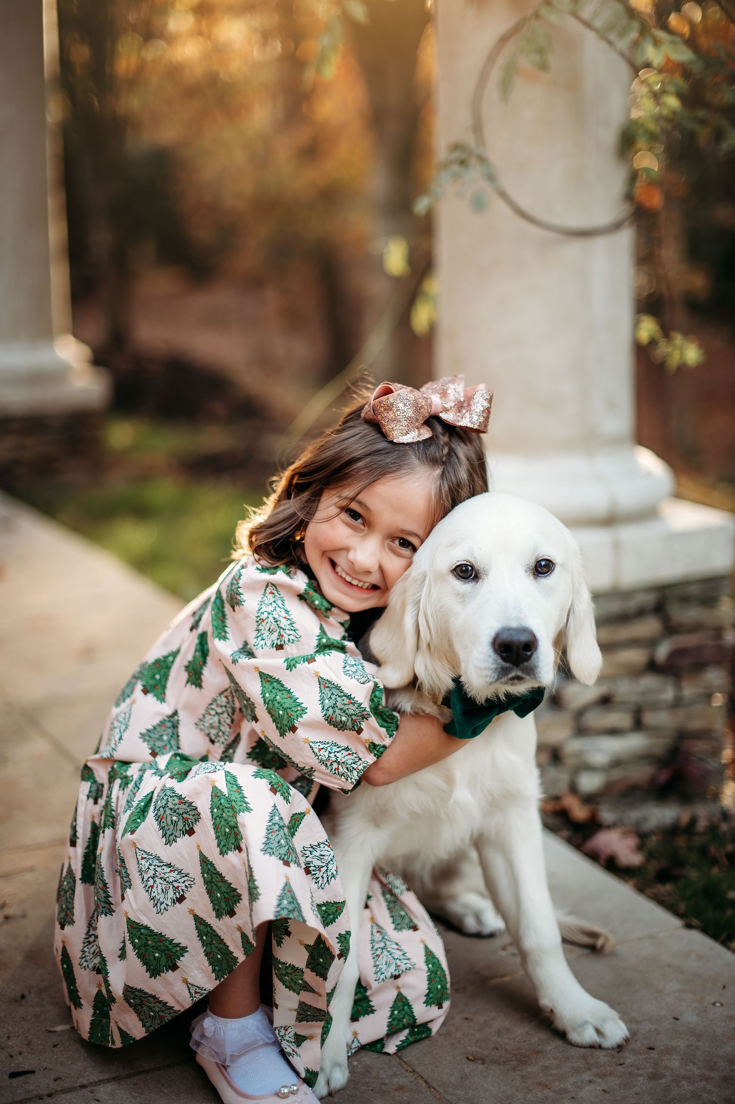 A young person in a tree-print dress hugs a white retriever dog while sitting on outdoor stone steps in warm lighting.