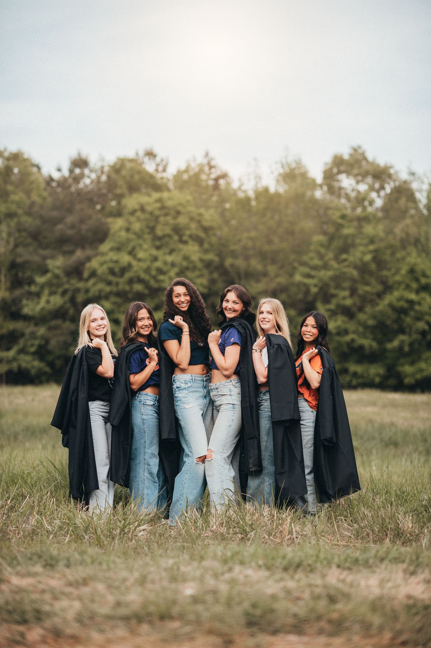 Six women in matching outfits pose together wearing black tops and jeans in a sunny field.