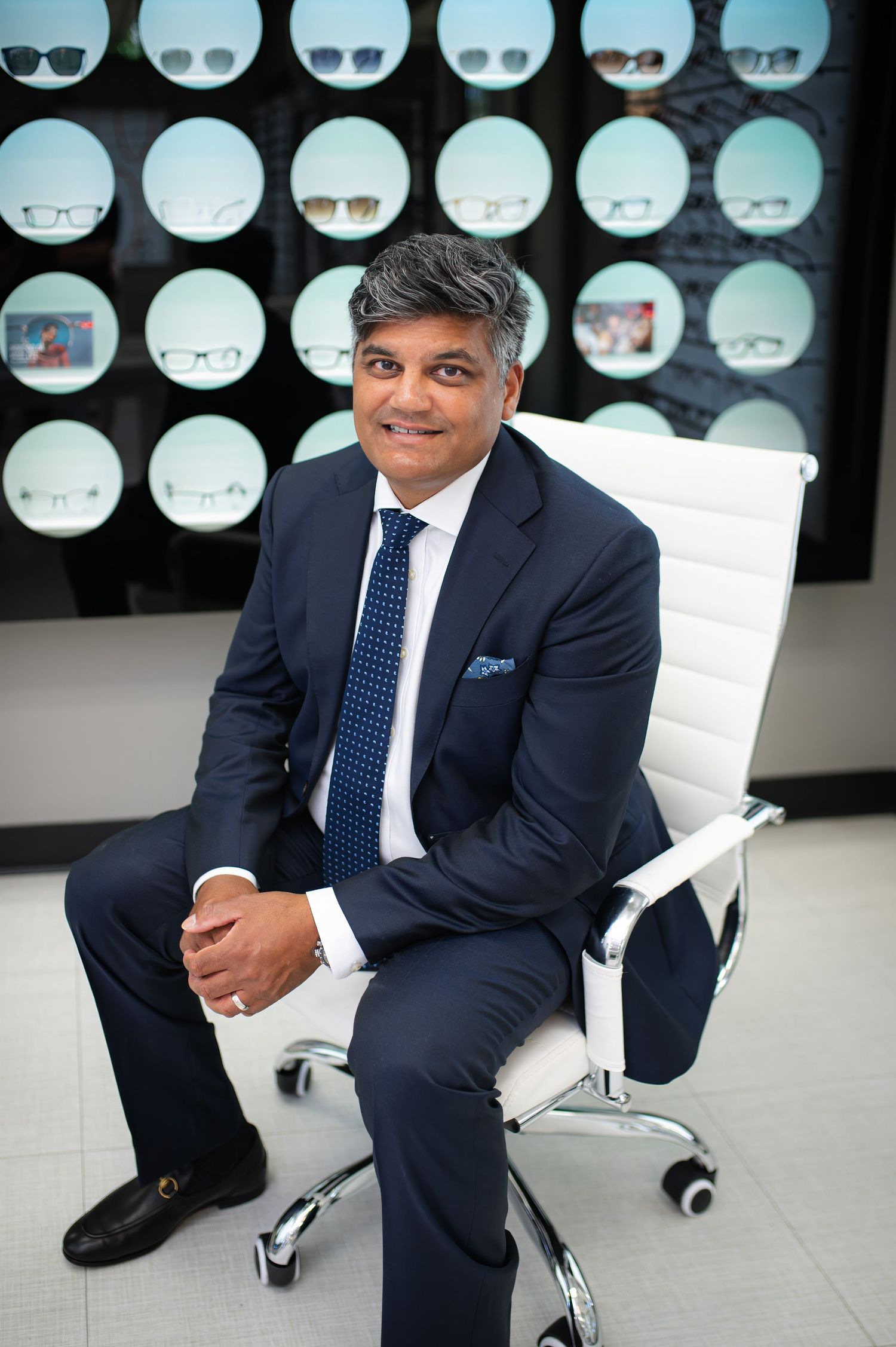 A professional in a navy suit sits in a modern white office chair with circular displays mounted on the wall behind.