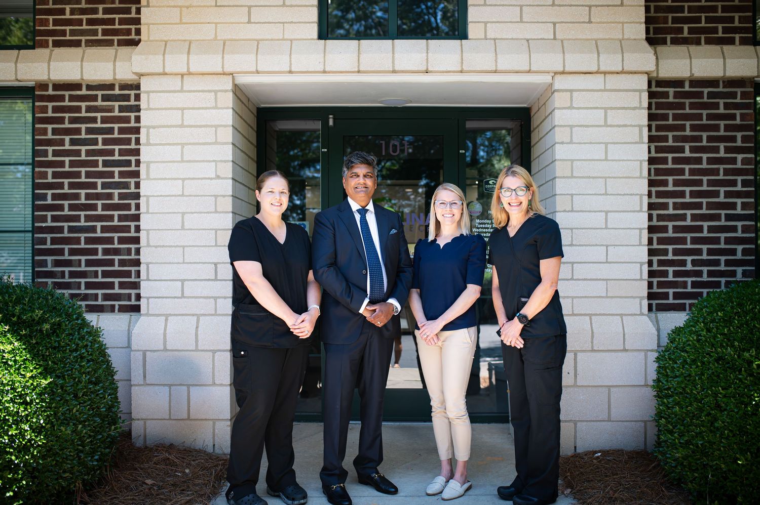 Professional business team standing together outside a brick building entrance with rounded arches and manicured shrubs.