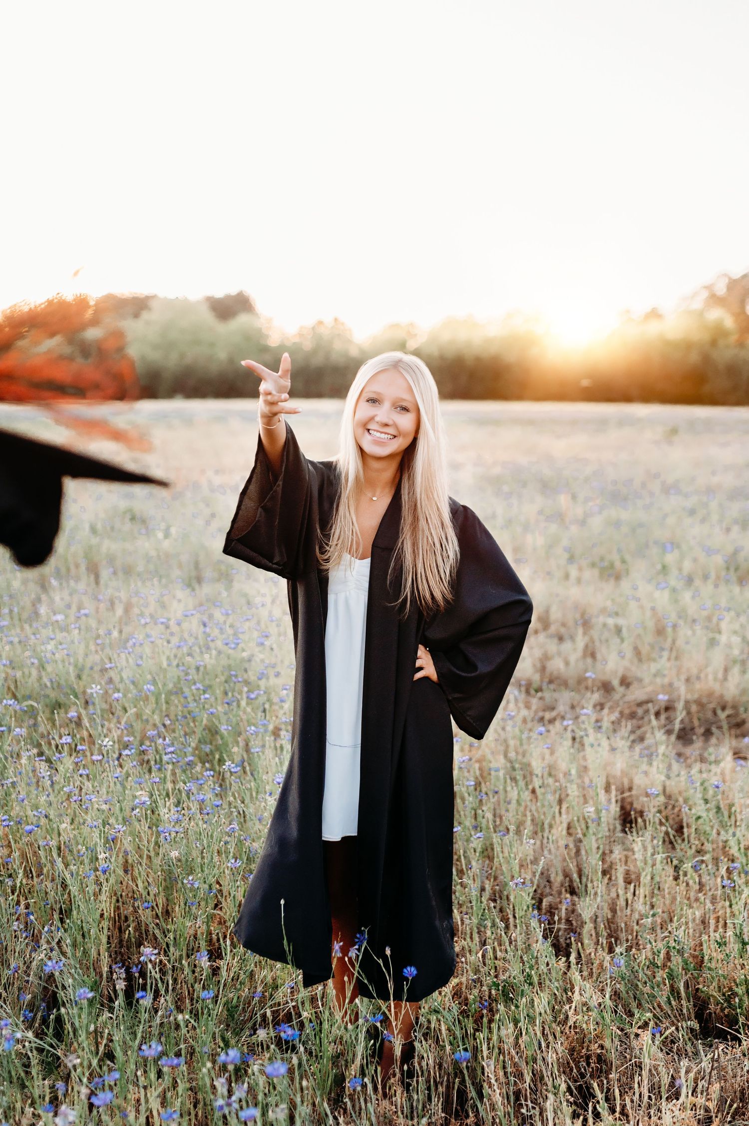 Smiling graduate in black gown tossing cap in golden field at sunset, long blonde hair, white dress underneath.