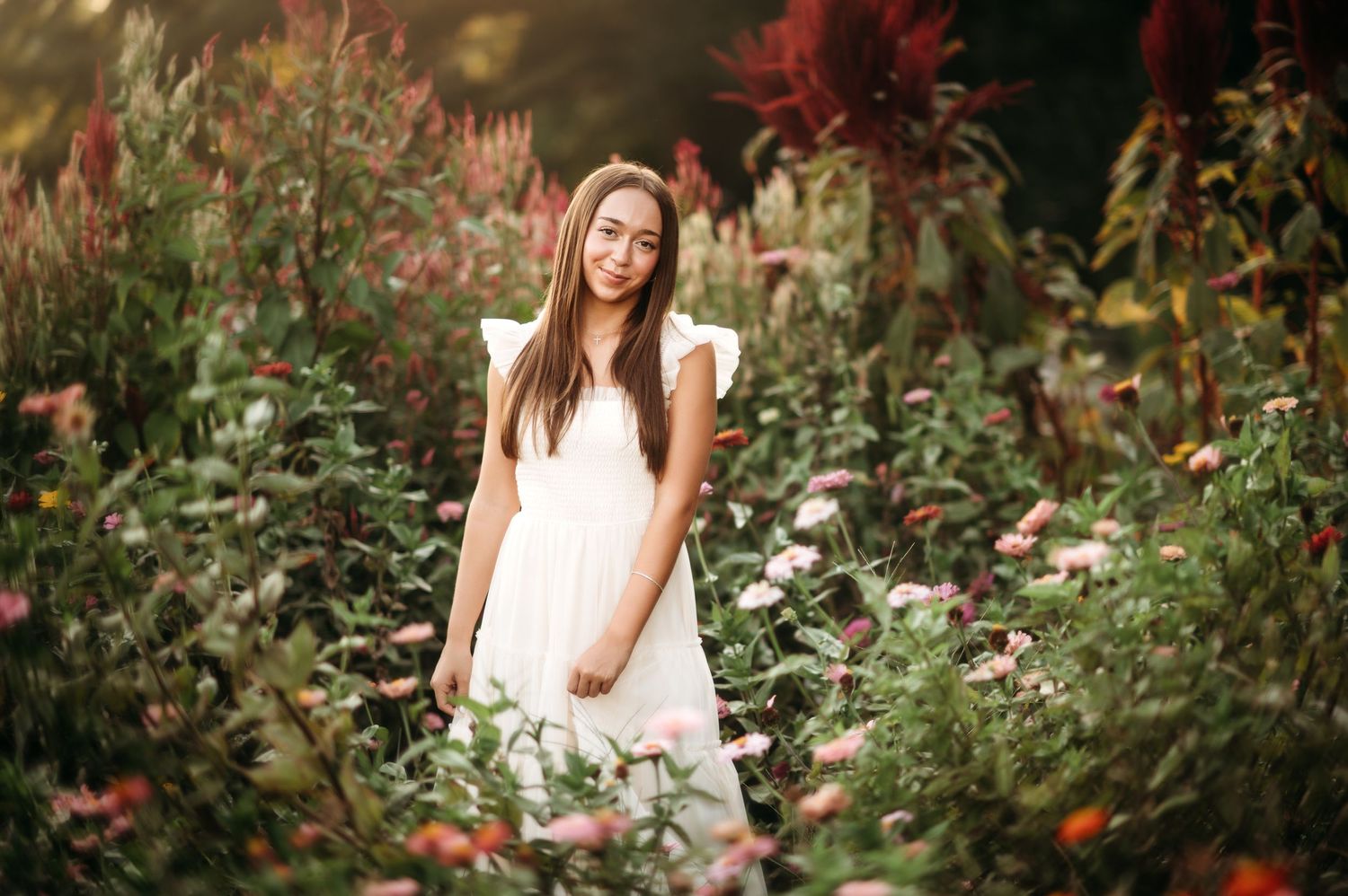 A person in a white dress smiles while standing in a vibrant garden filled with colorful flowers.