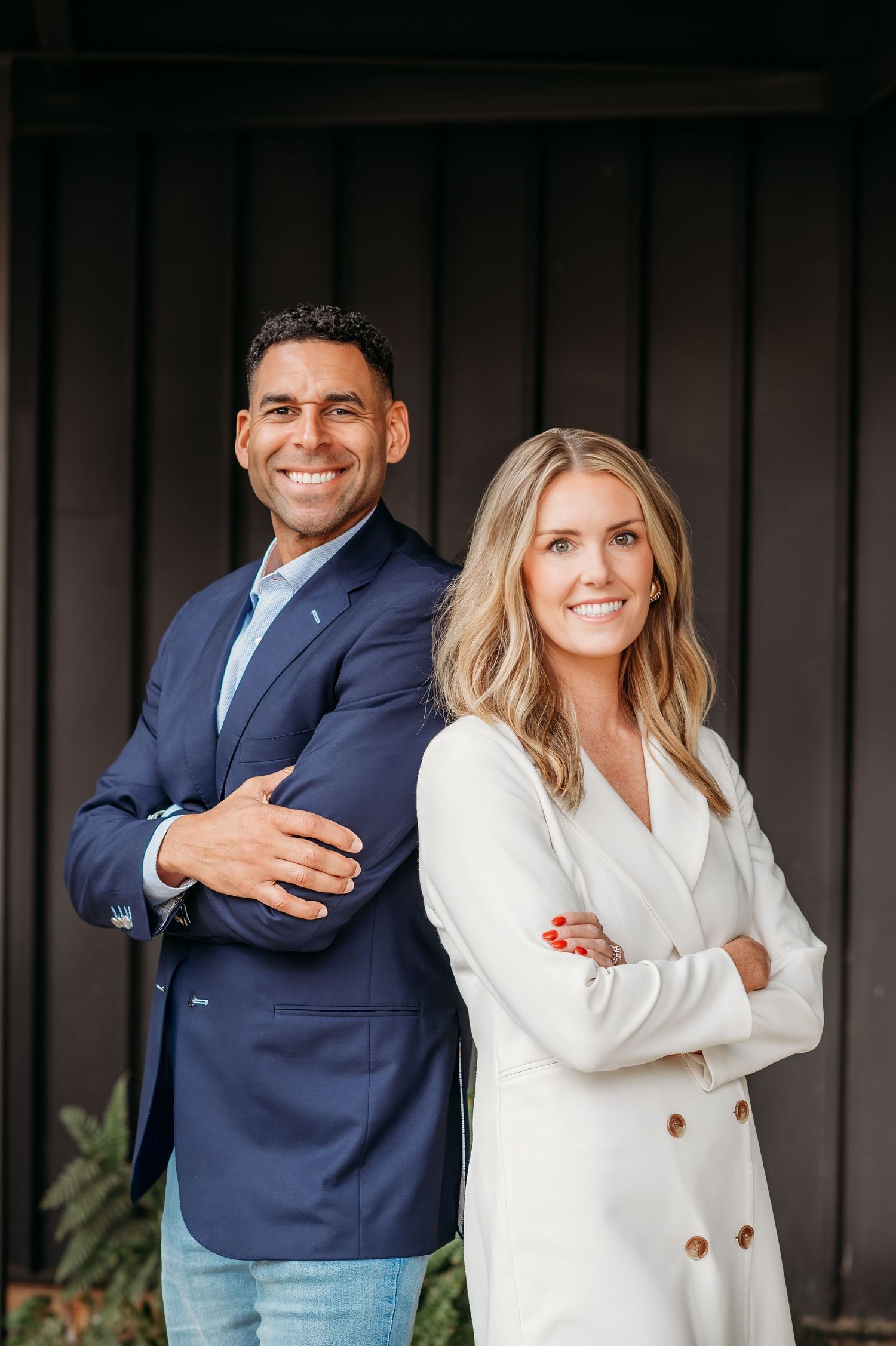 Professional business portrait of two colleagues in business attire standing back-to-back against dark wall outdoors.