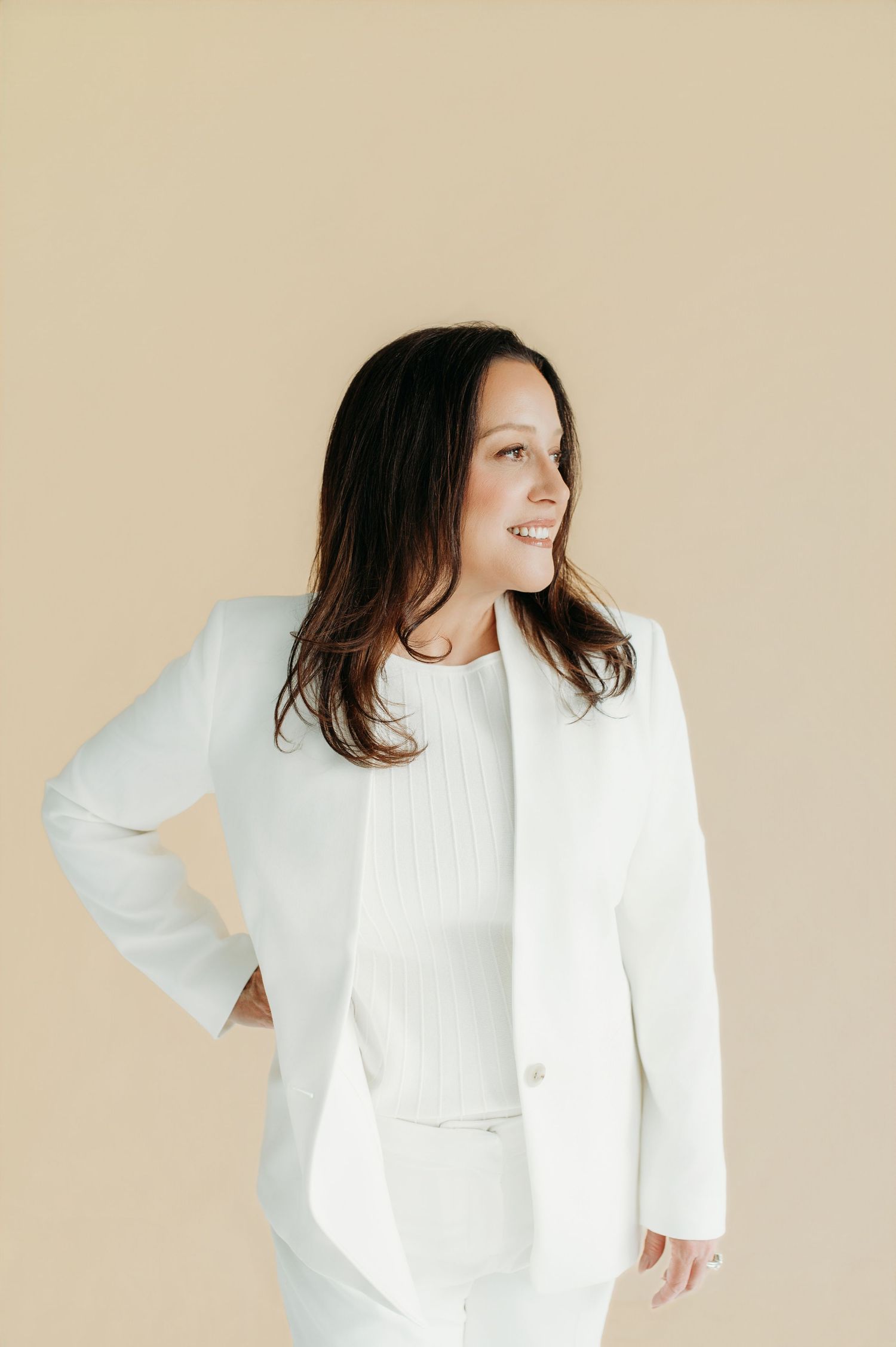 Smiling woman with dark hair wearing a white blazer and pants suit against a beige background.