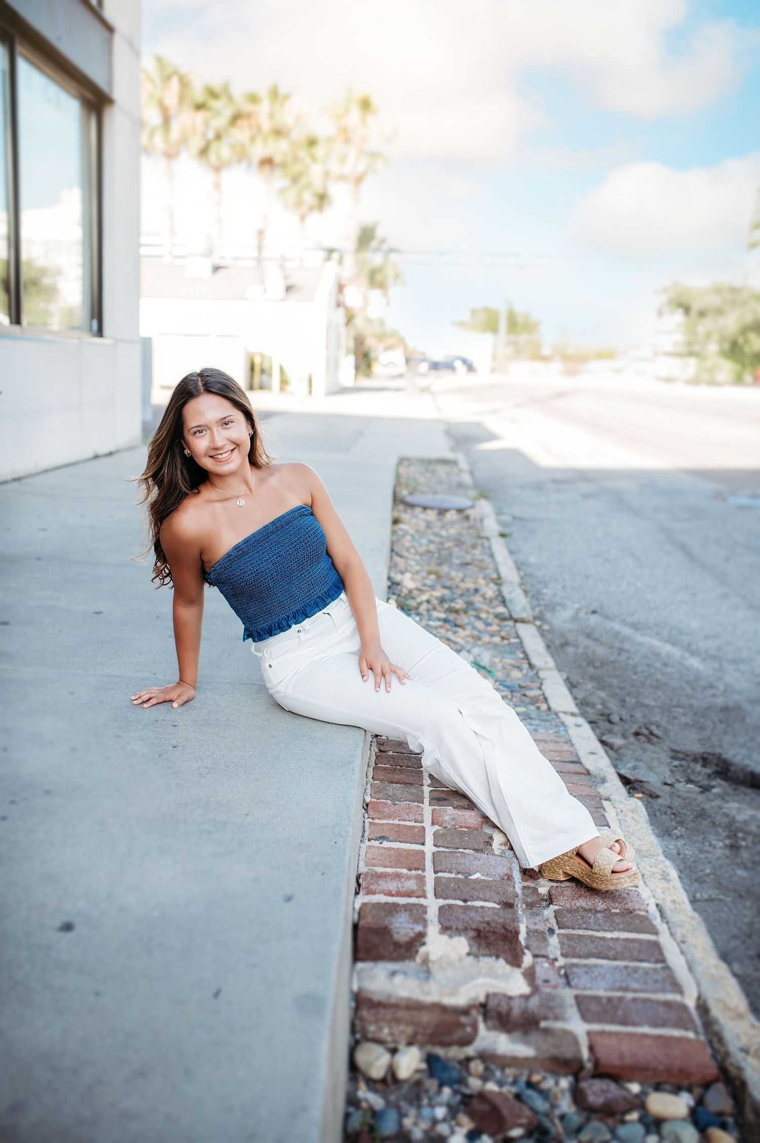 Casual summer portrait on brick ledge wearing denim and blue top in bright sunlight.