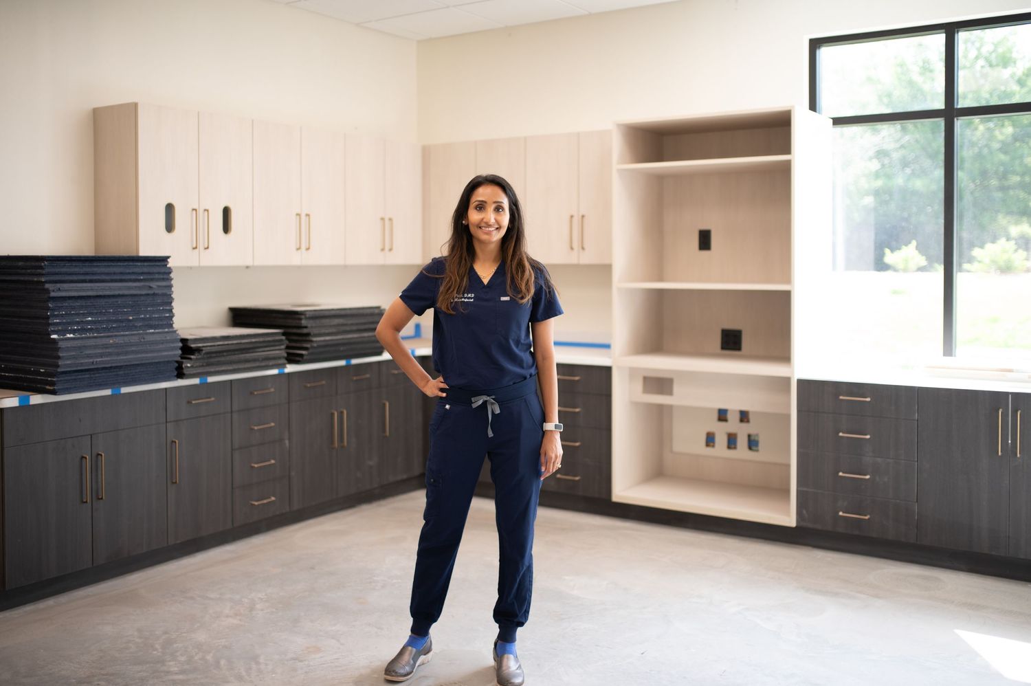 Healthcare professional stands confidently in modern medical office with dark cabinets and bright window lighting.
