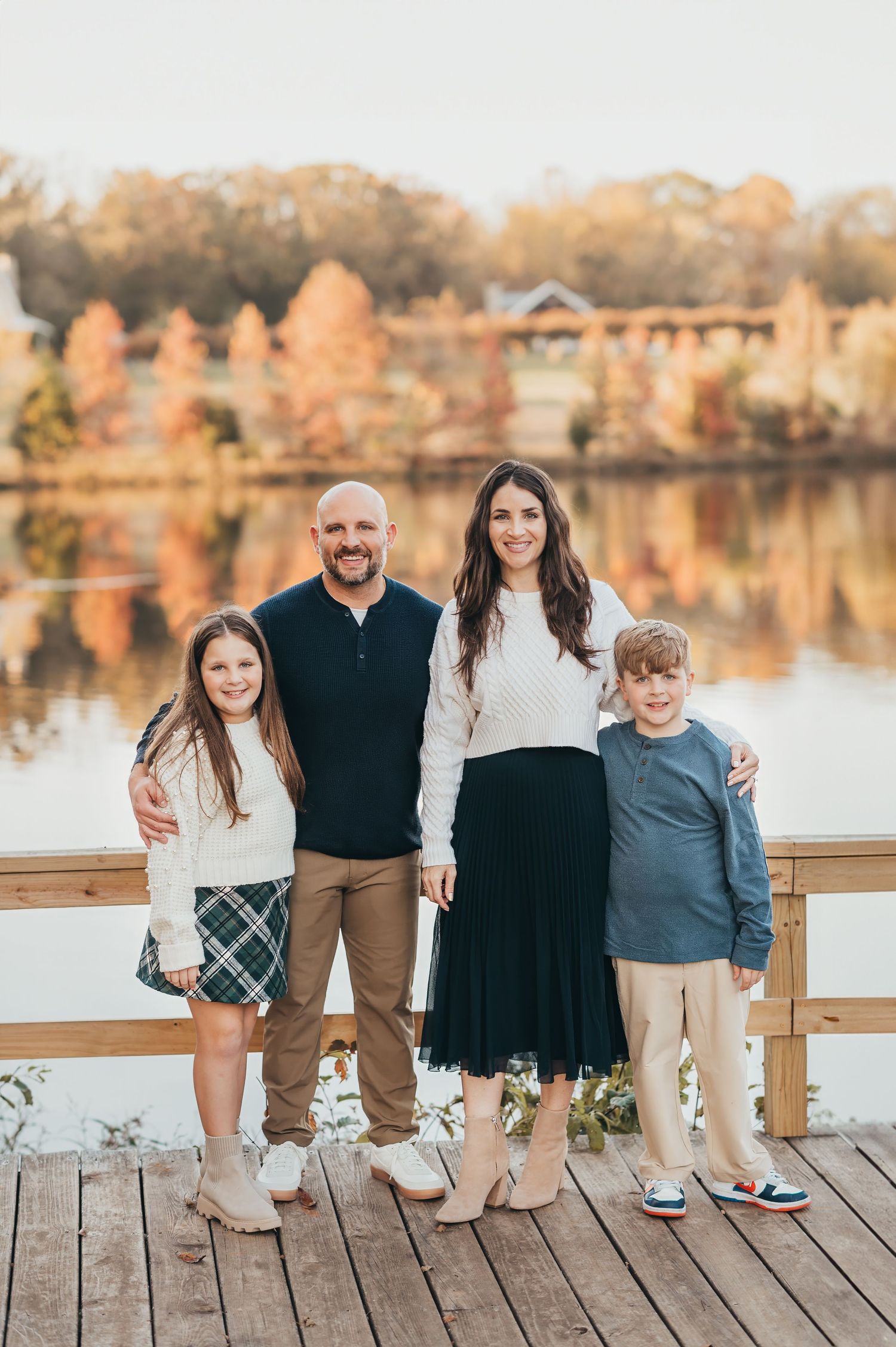 Family of four poses together on wooden deck by autumn lake with colorful fall foliage reflection in background.