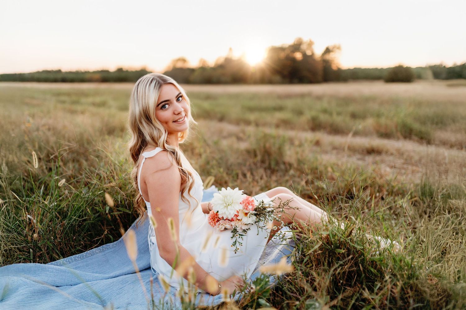 A person in a white dress sits on a blue blanket in a sunlit field at golden hour.