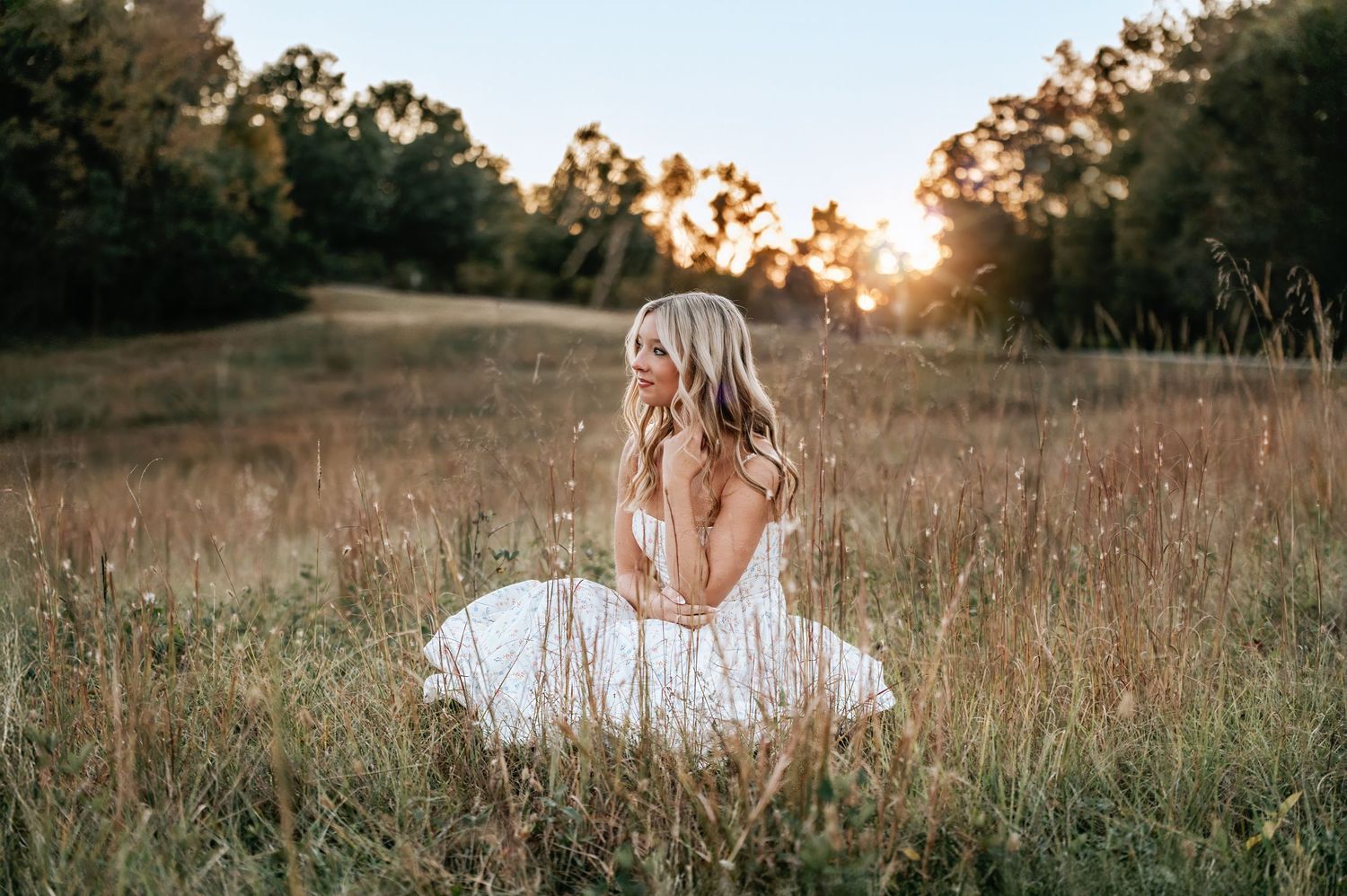 A woman in a white dress sits in a field at sunset, surrounded by tall grass and trees. High school senior portrait session Fort Mill SC