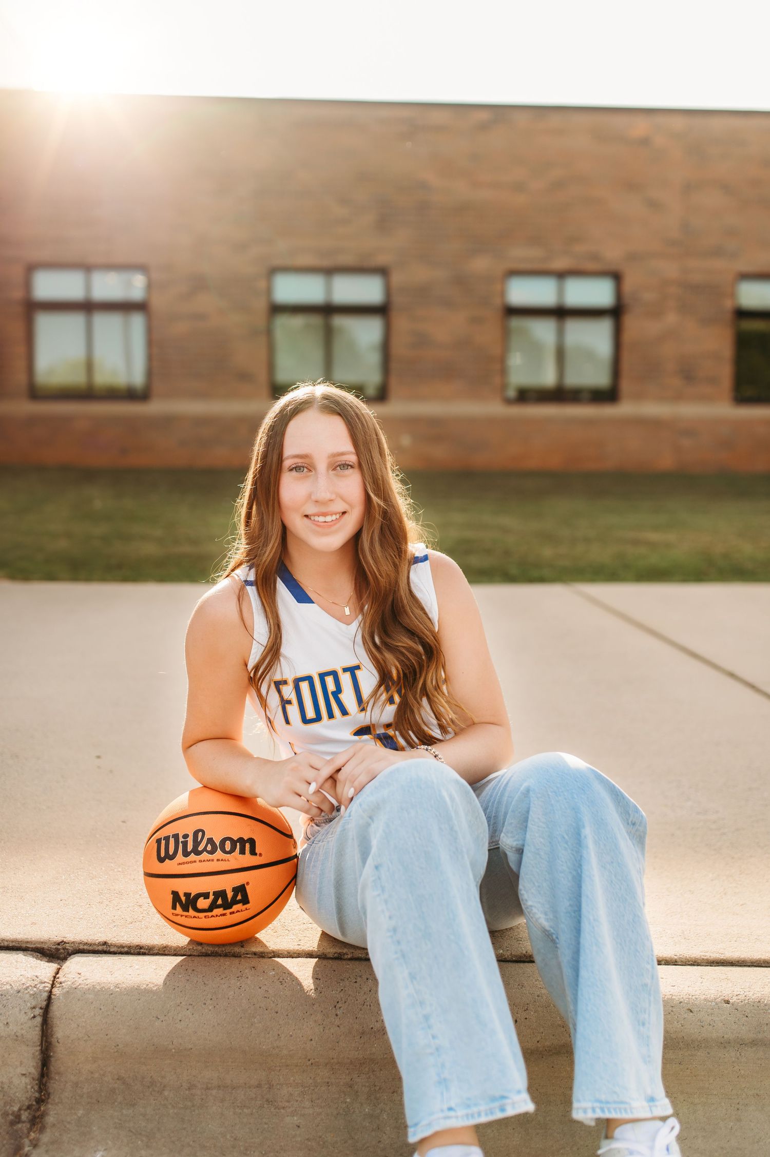 Student athlete sits with basketball wearing Fort Mill jersey and jeans outside brick building.