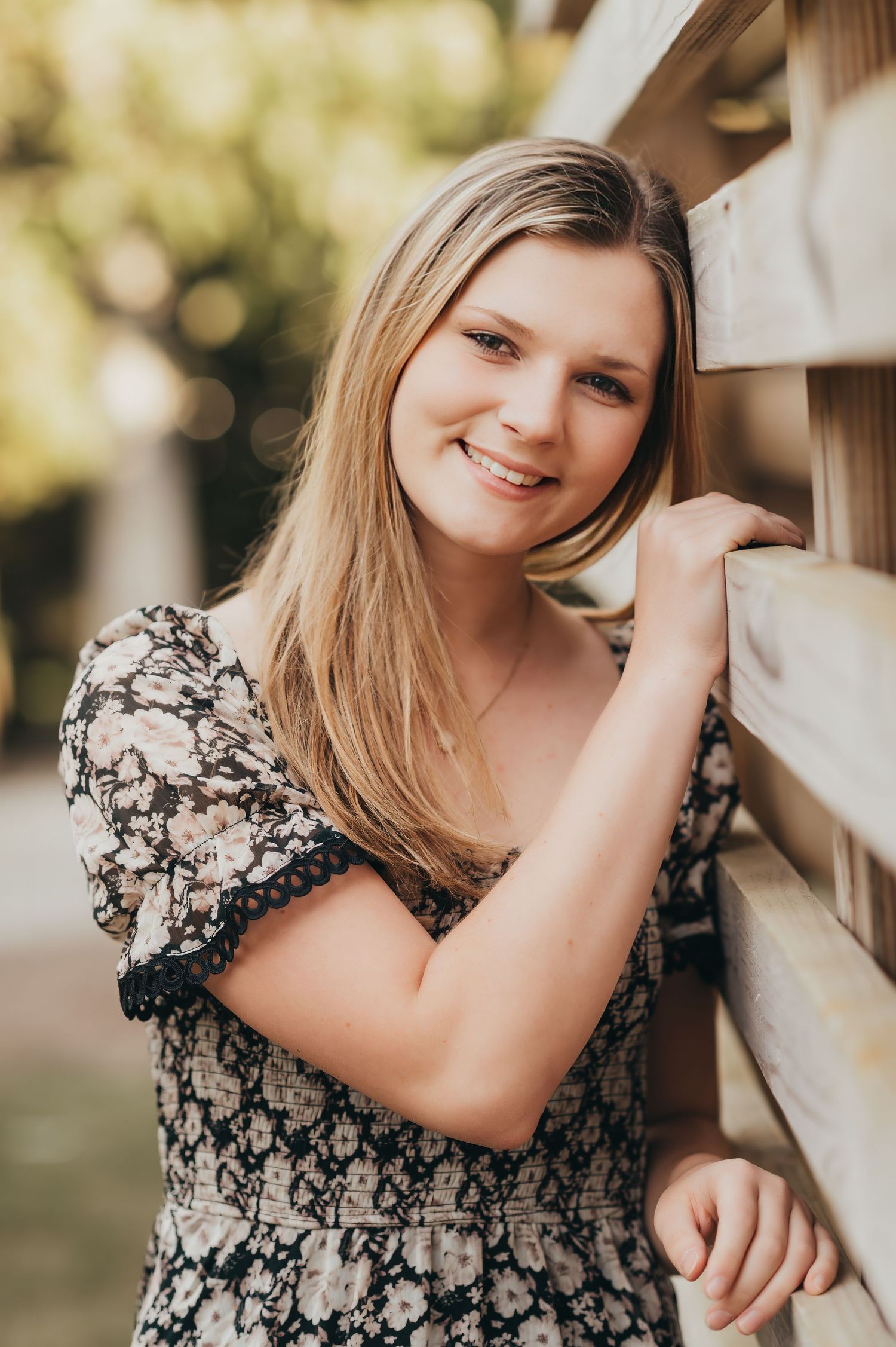 Portrait against wooden fence wearing a floral patterned dress in warm natural lighting.