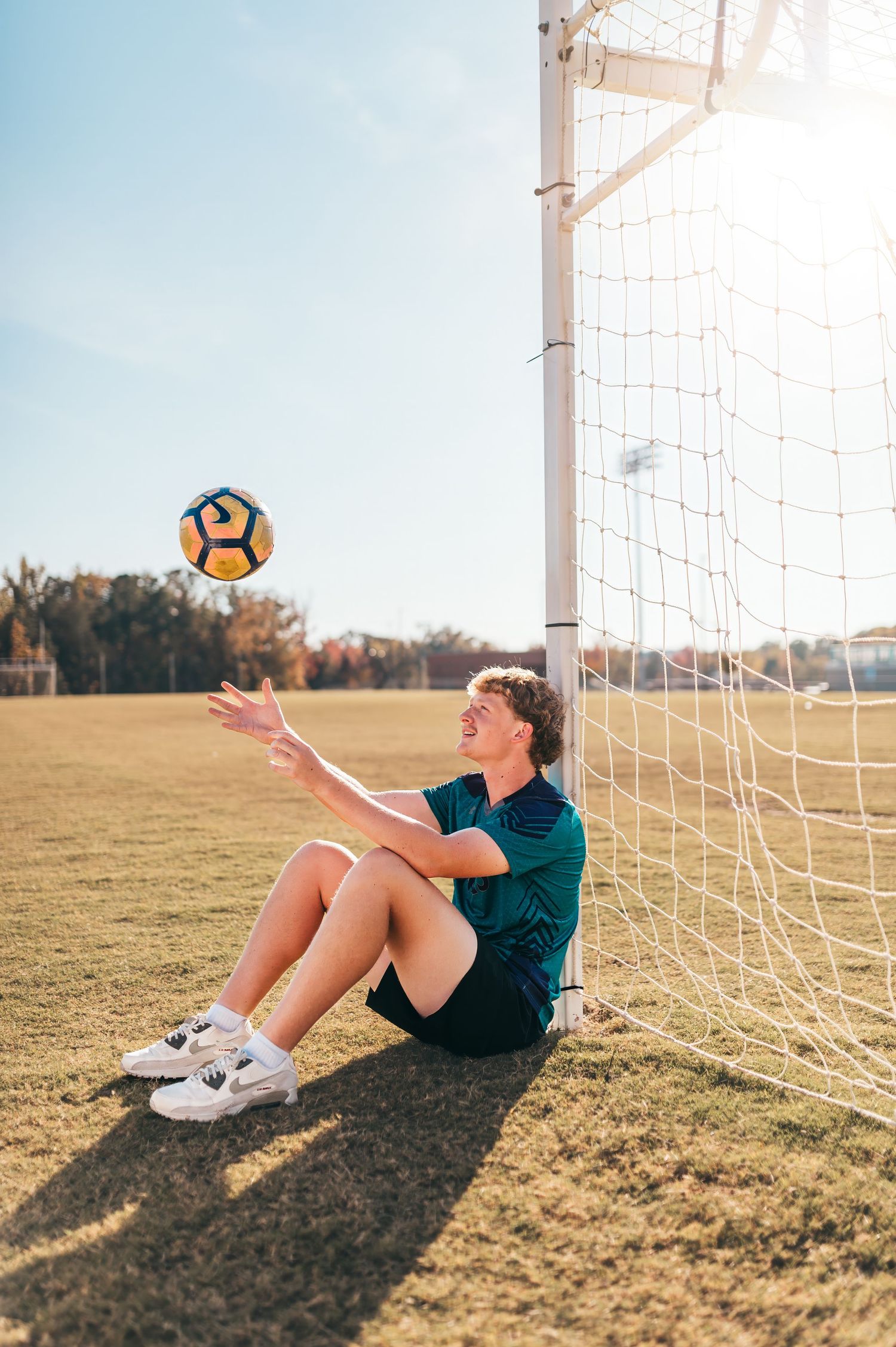 Soccer player resting by goal post with ball during golden hour practice.