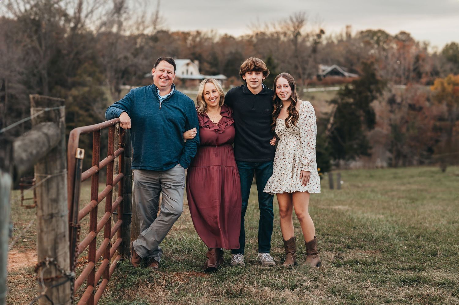 Group standing together and leaning against rustic metal gate in autumn field with houses in background at sunset.