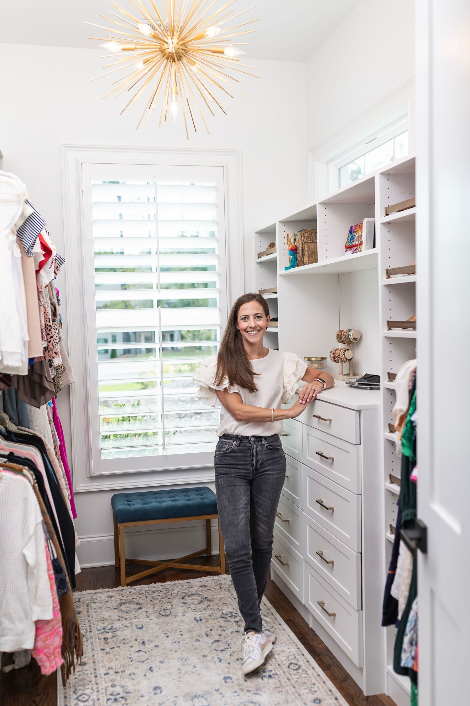 Bright modern walk-in closet with built-in white shelving, gold starburst light fixture, and patterned area rug on floor.