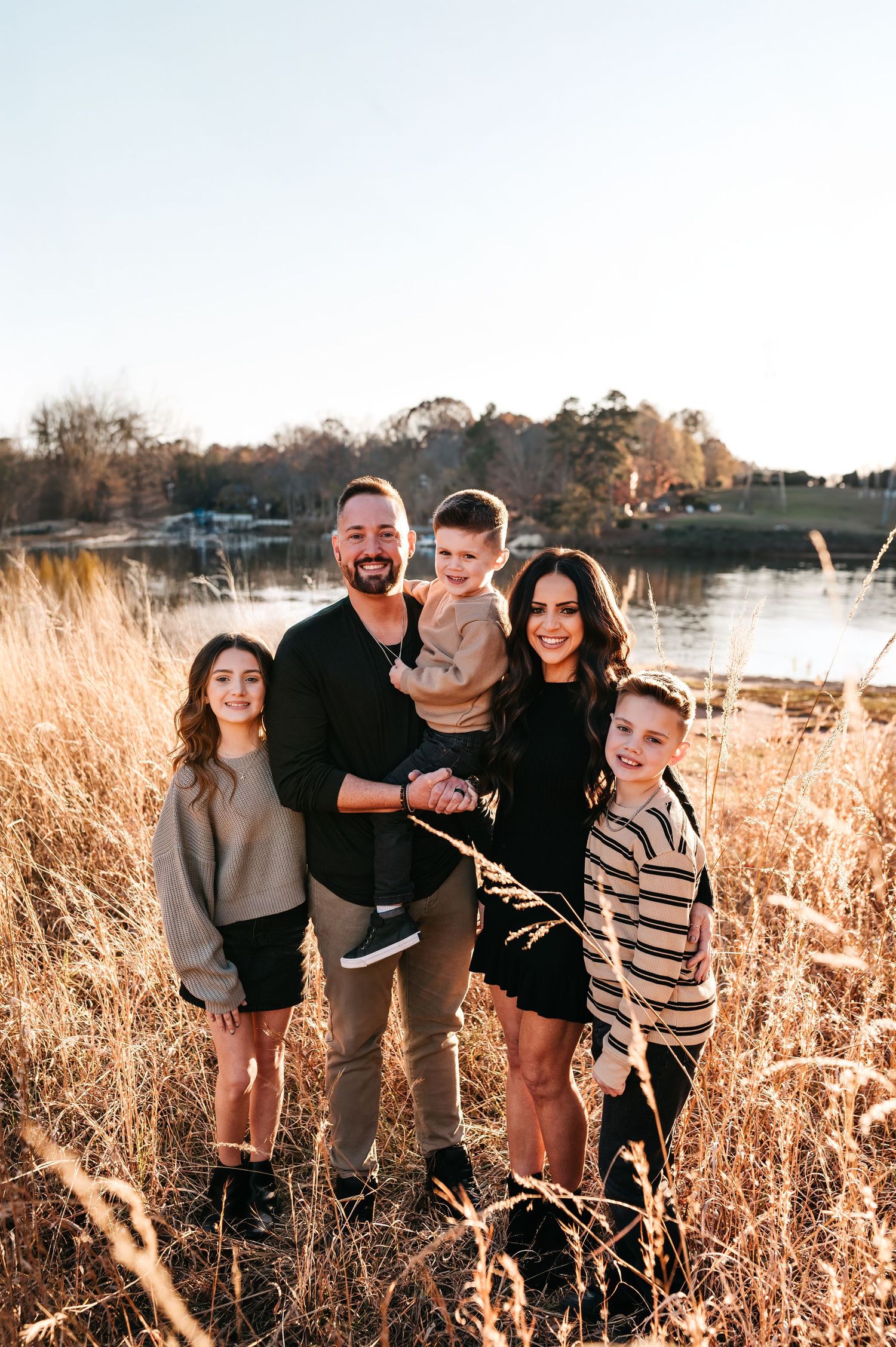A group stands together in tall golden grass at sunset near a lake, creating a warm autumn family portrait scene.