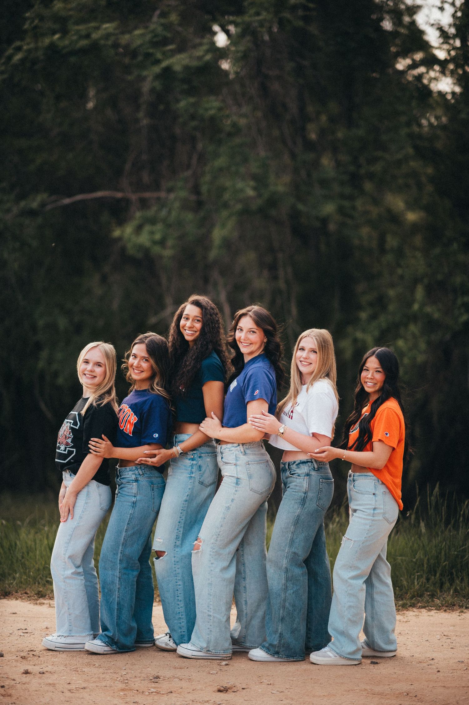 Group of six friends in coordinated denim jeans pose together outdoors on a dirt path.