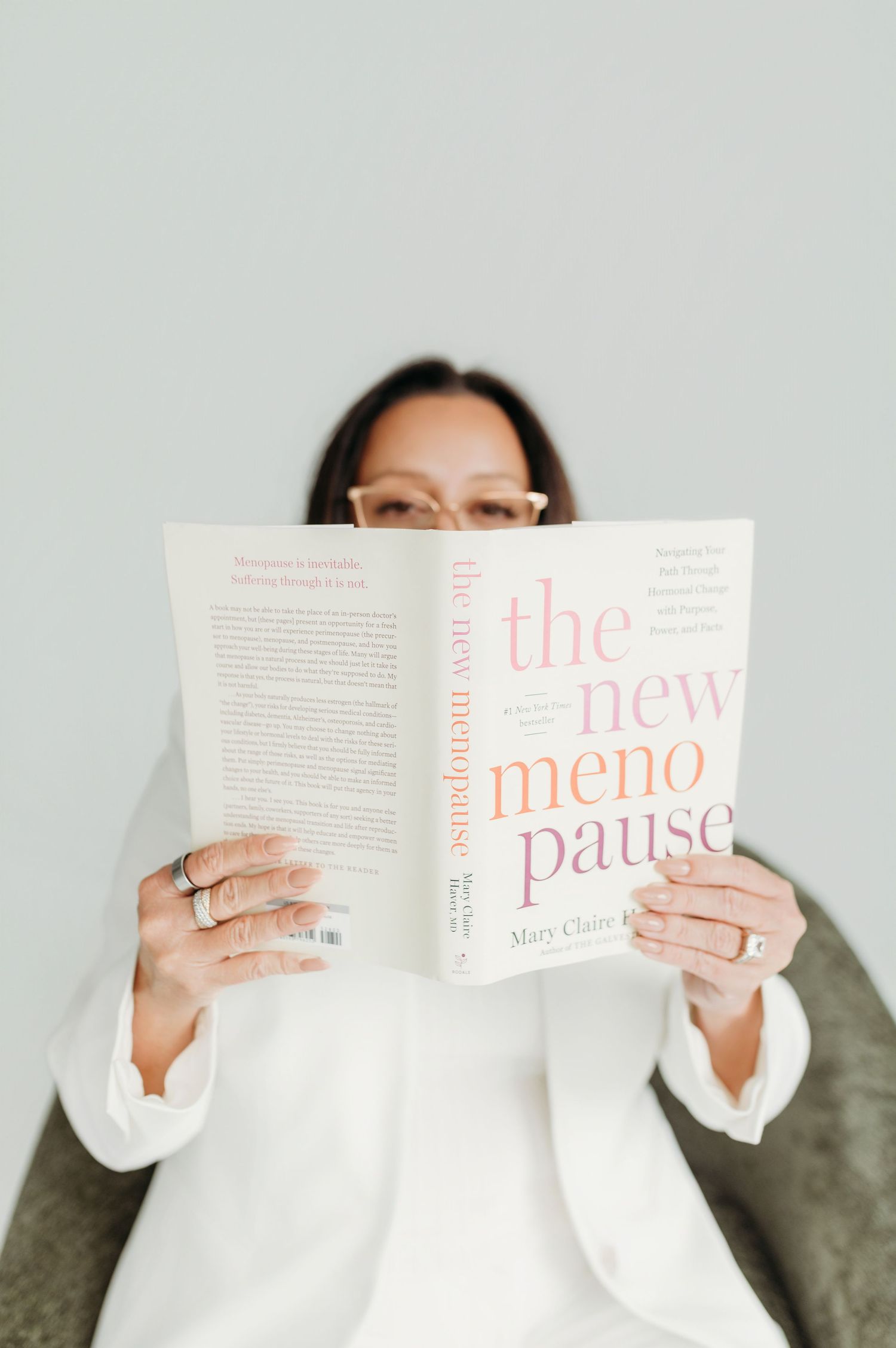Woman in white blazer reading 'The New Menopause' by Mary Claire, seated against a light gray background.