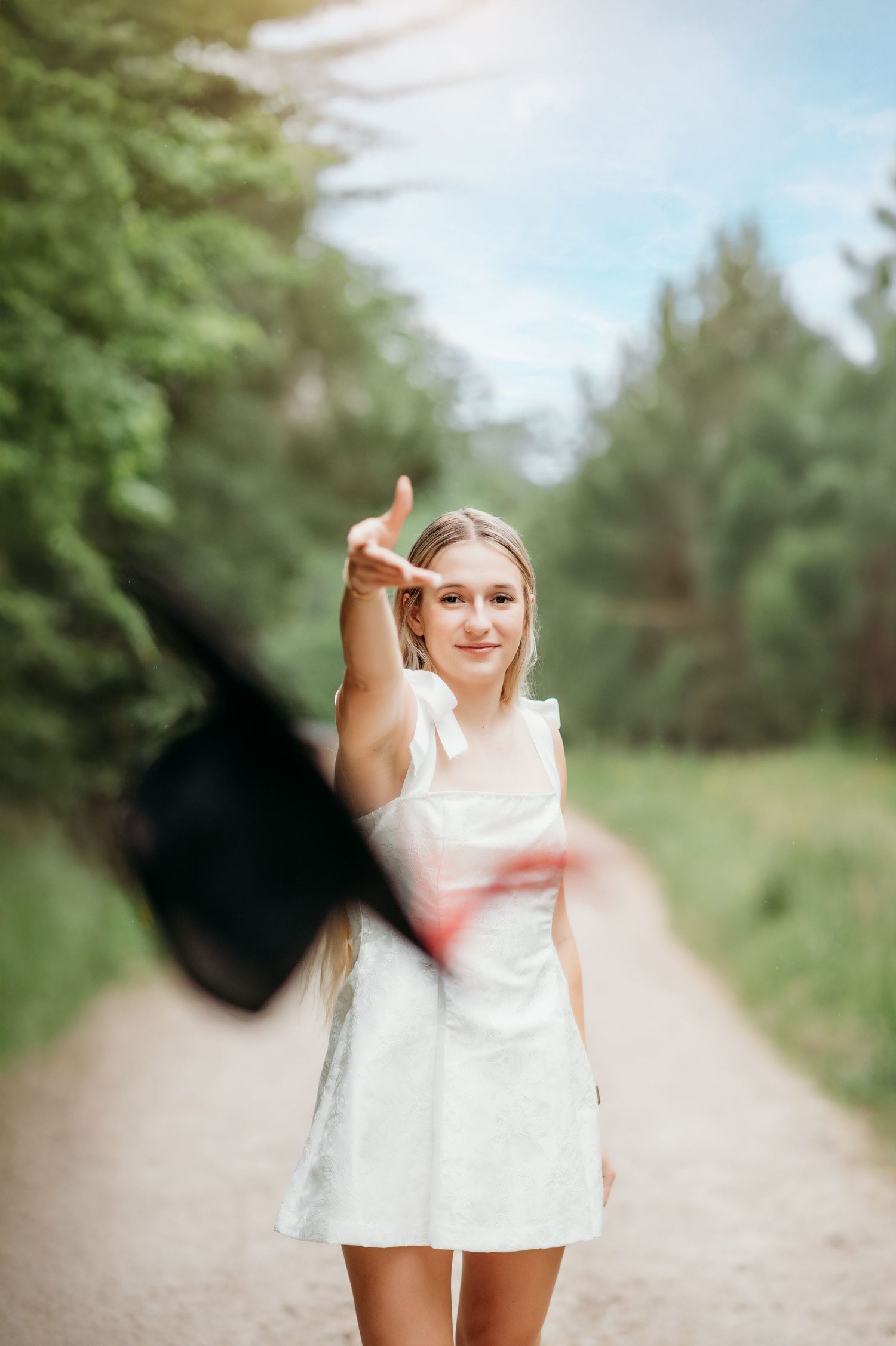 A graduate in white dress tosses their cap on a tree-lined path during a celebration photoshoot.