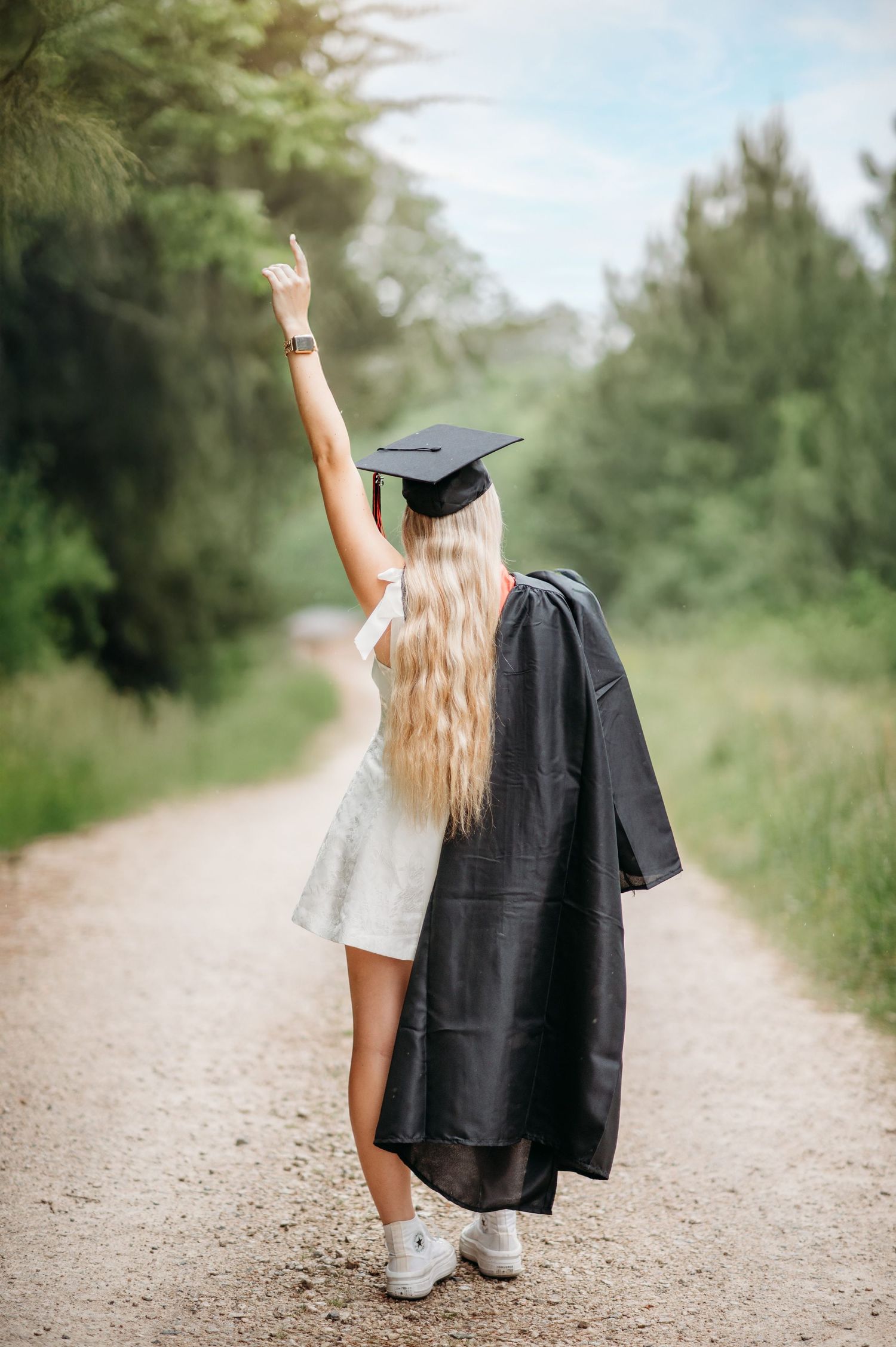 A graduate in cap and gown raises their arm triumphantly on a dirt path surrounded by trees.