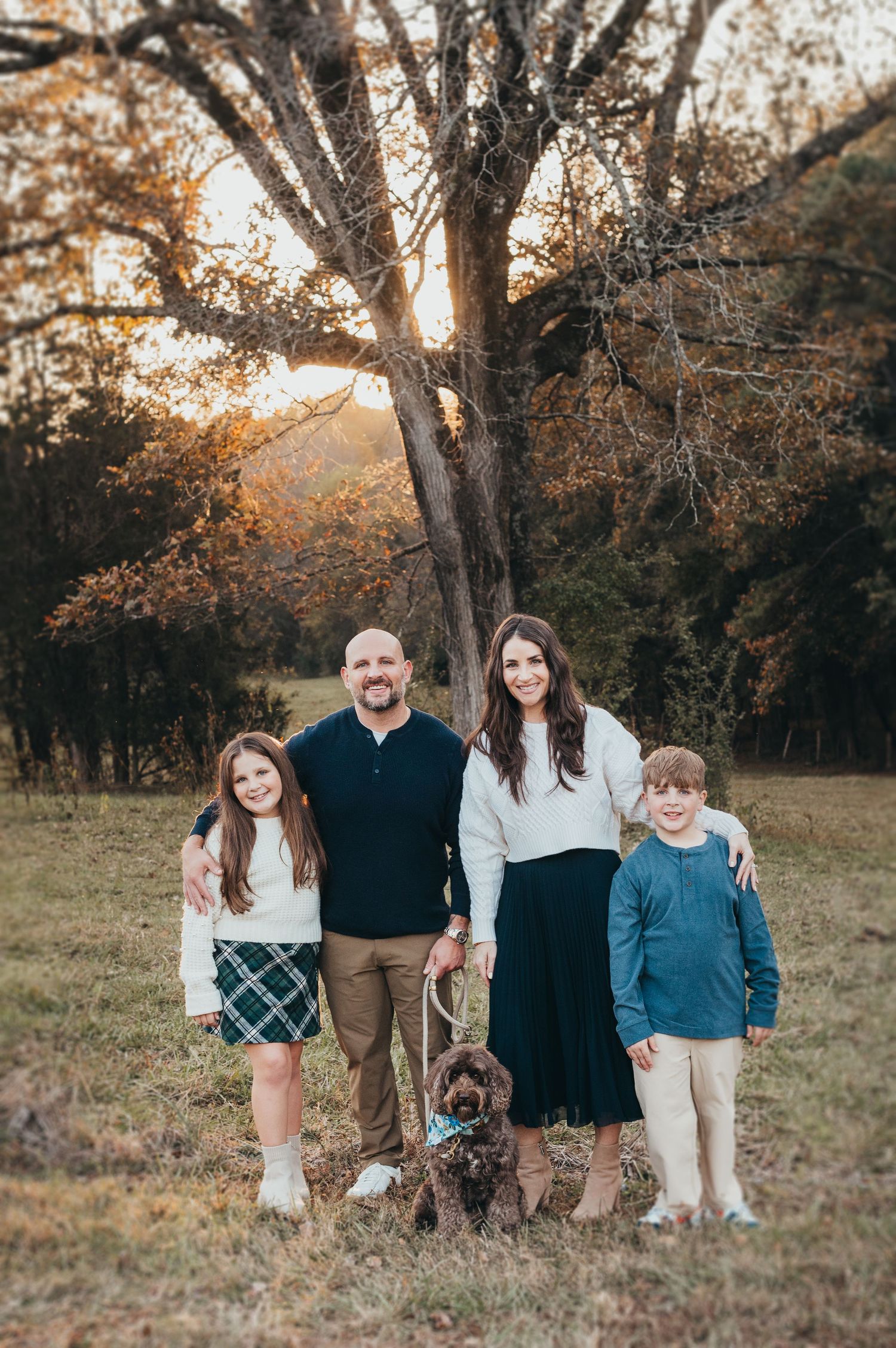 A family poses together with their dog during a fall sunset in a wooded field with warm autumn colors.
