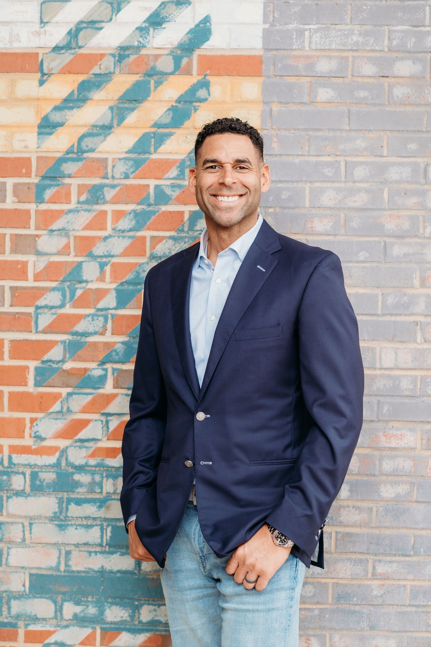 Professional portrait of person in navy blazer and light blue dress shirt against brick wall with decorative painted pattern.