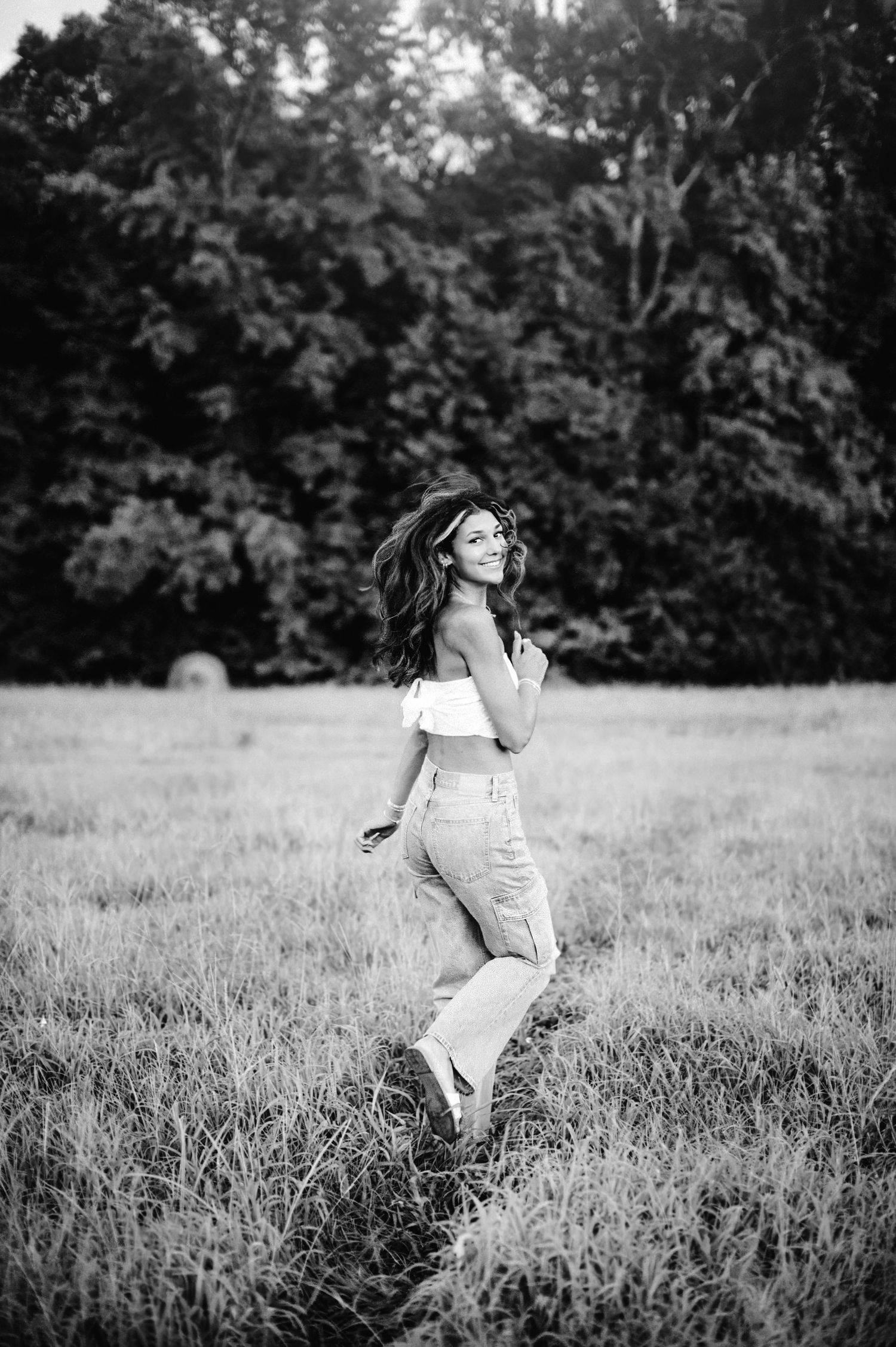 Black and white photo of someone in a white top and light pants twirling playfully in a grassy meadow at dusk.