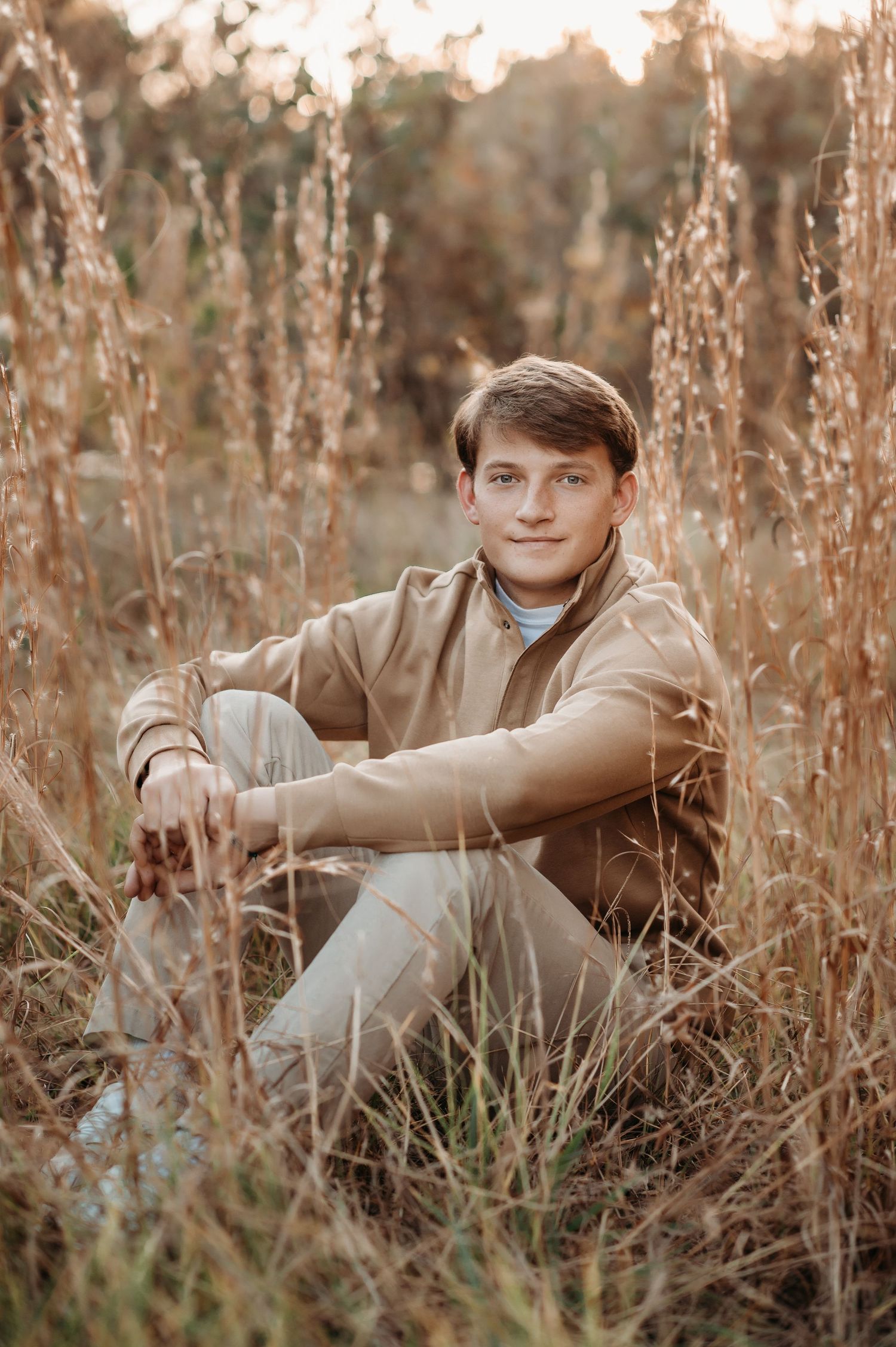 A young person in casual attire sits among tall grass during a golden hour photoshoot.