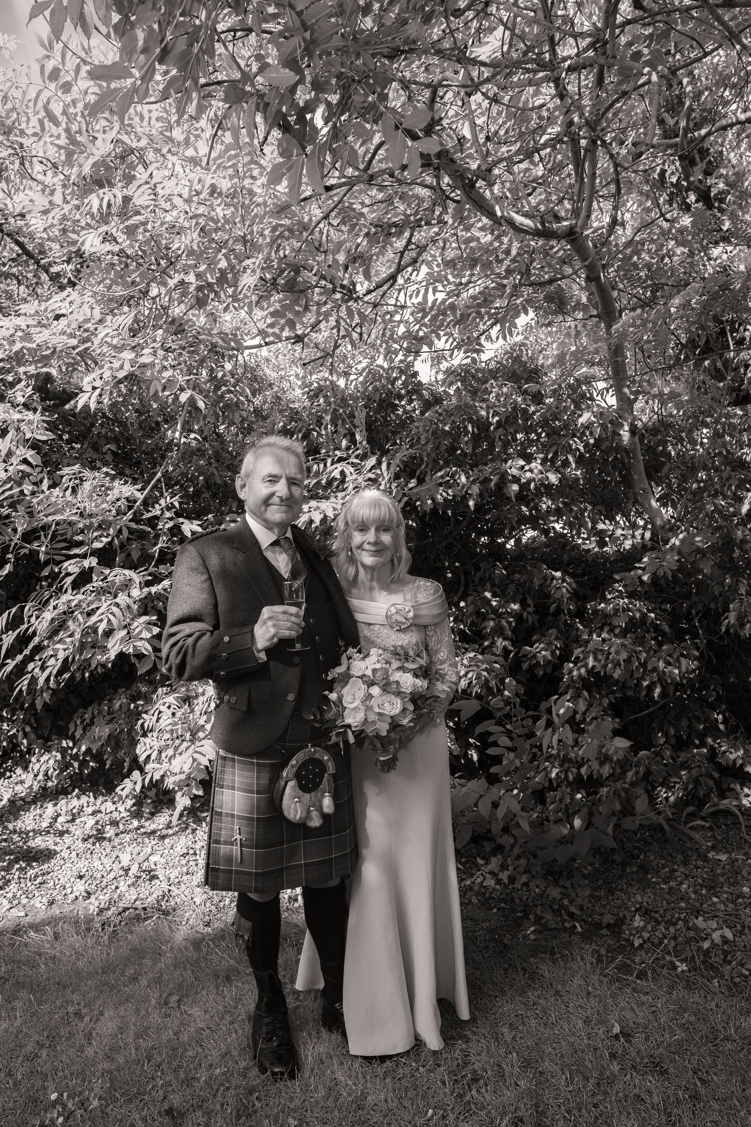 A black and white wedding photo showing a couple in formal attire with traditional Scottish kilt outdoors among foliage.