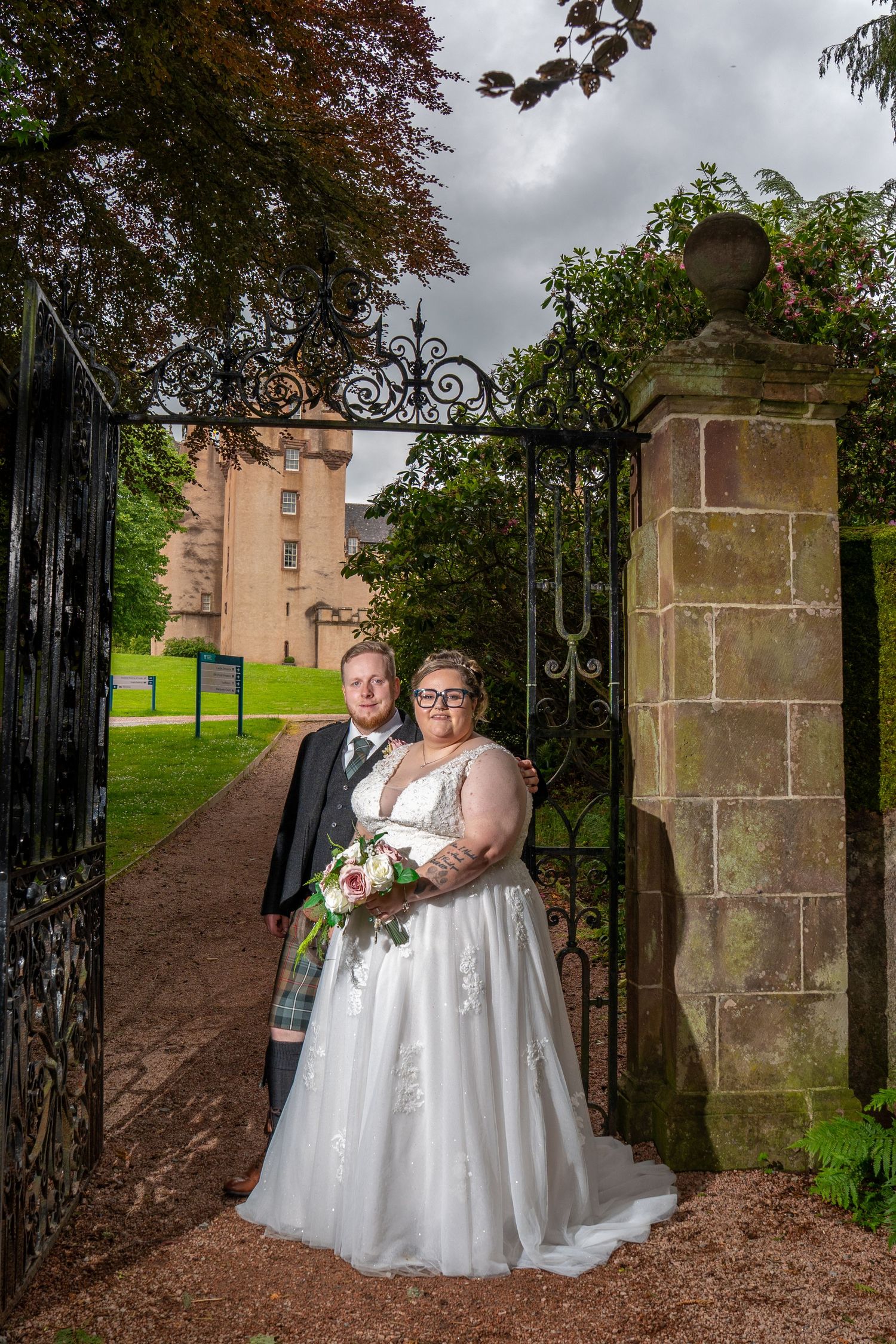 A couple in matching white wedding dresses pose together in front of a stone wall and ornate black iron gate on their special day.