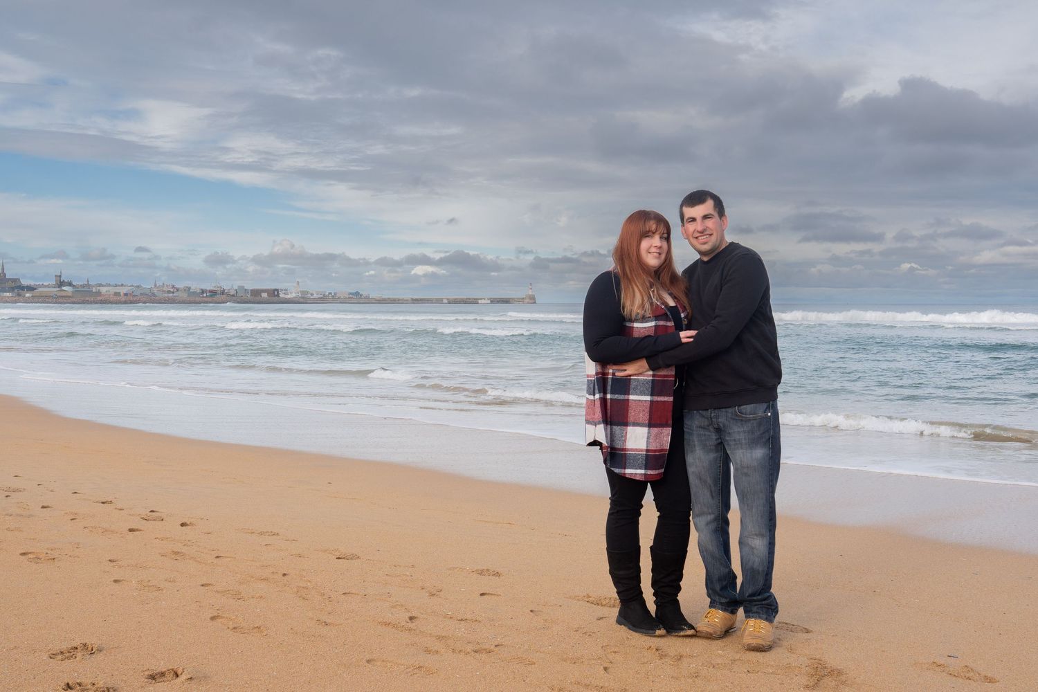 A couple standing together on a sandy beach with cloudy skies and ocean waves in the background.