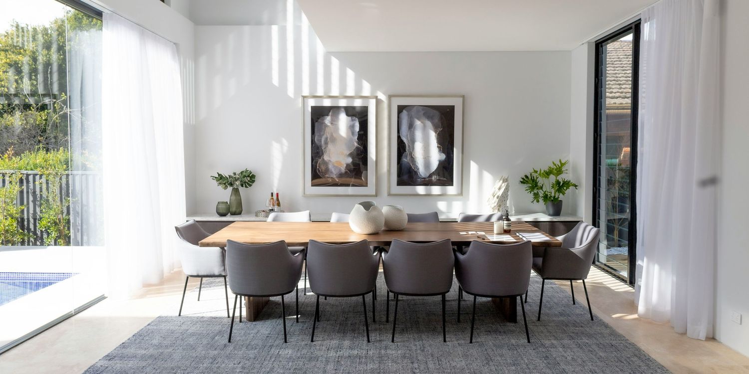Modern dining room with a long wooden table, upholstered gray chairs, black and white artwork, and natural lighting.