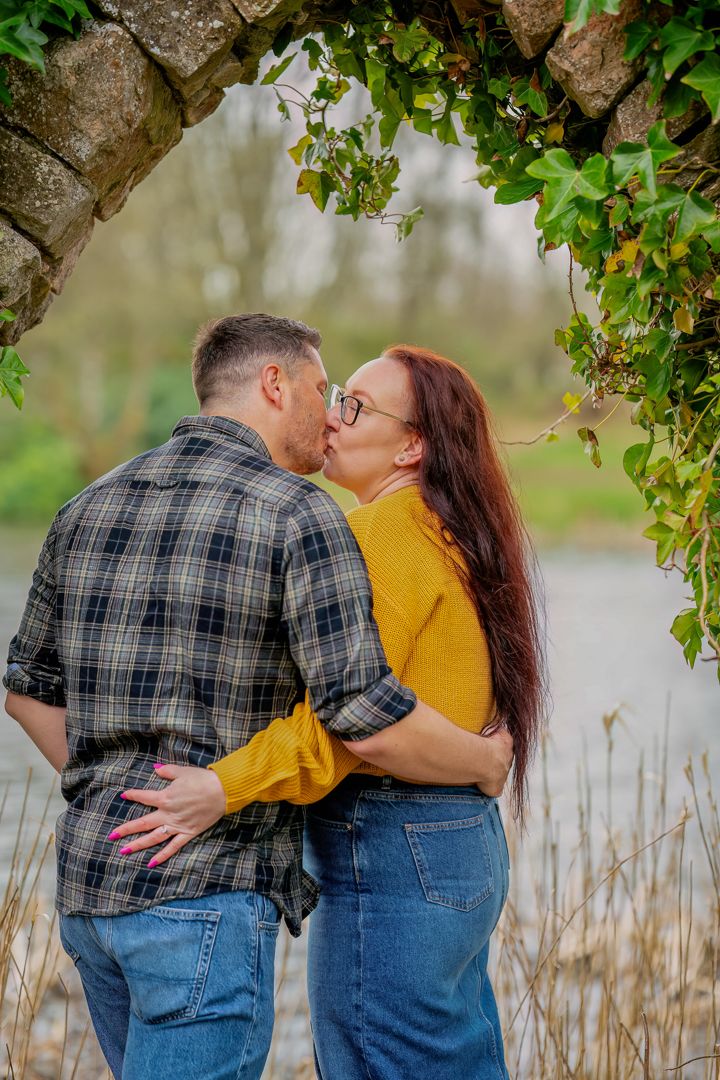 A romantic couple in denim and plaid share tender moments by a lake, with green foliage framing their embrace.