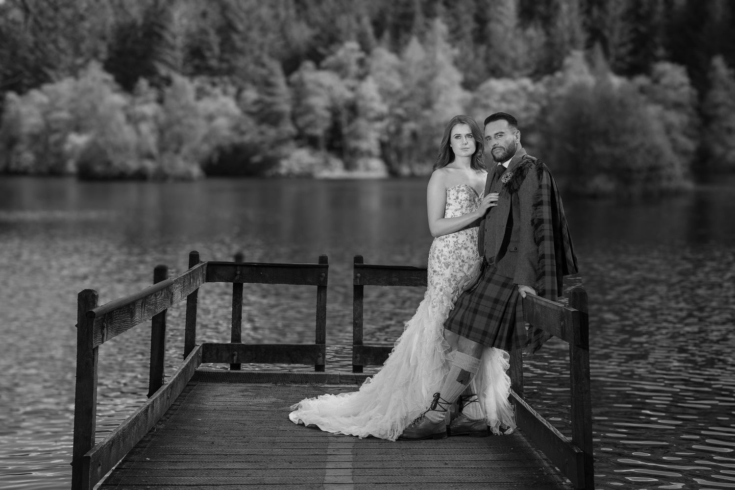 A romantic wedding couple sits together on a wooden dock overlooking a serene lake surrounded by trees.