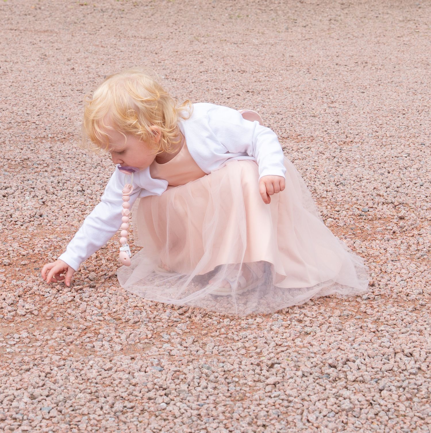 Small child in a white dress bending down to touch the ground on a reddish dirt surface.