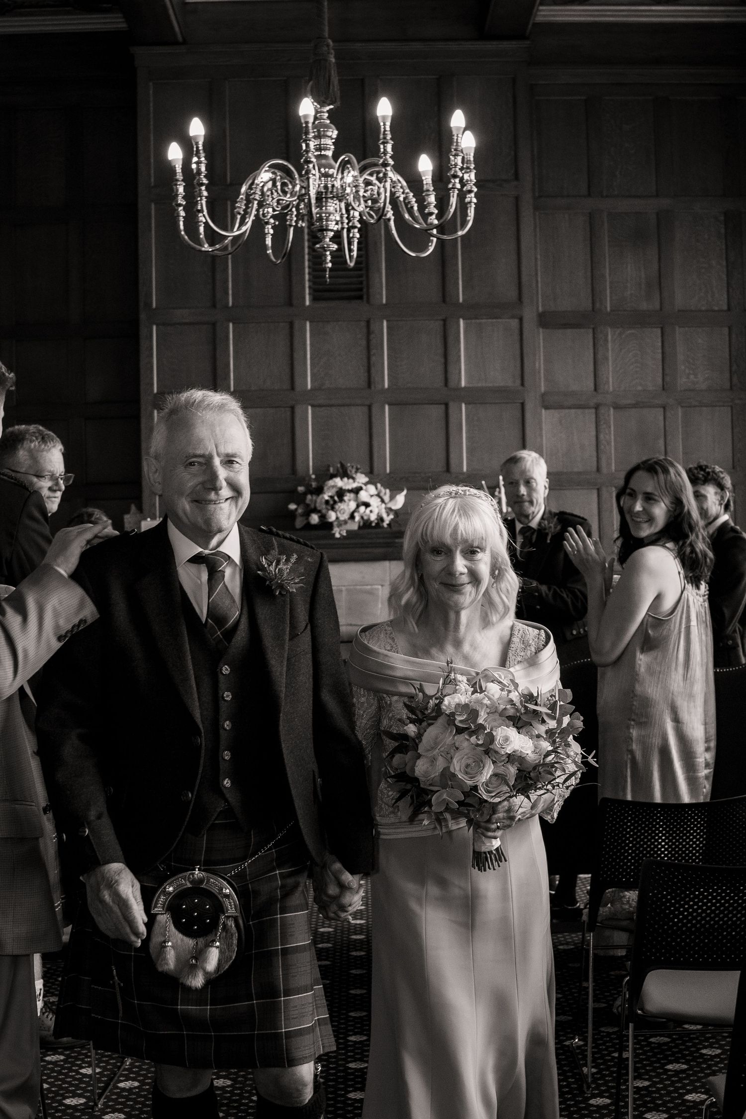 A black and white wedding photo showing a couple under an elegant chandelier in a formal setting with guests in background.