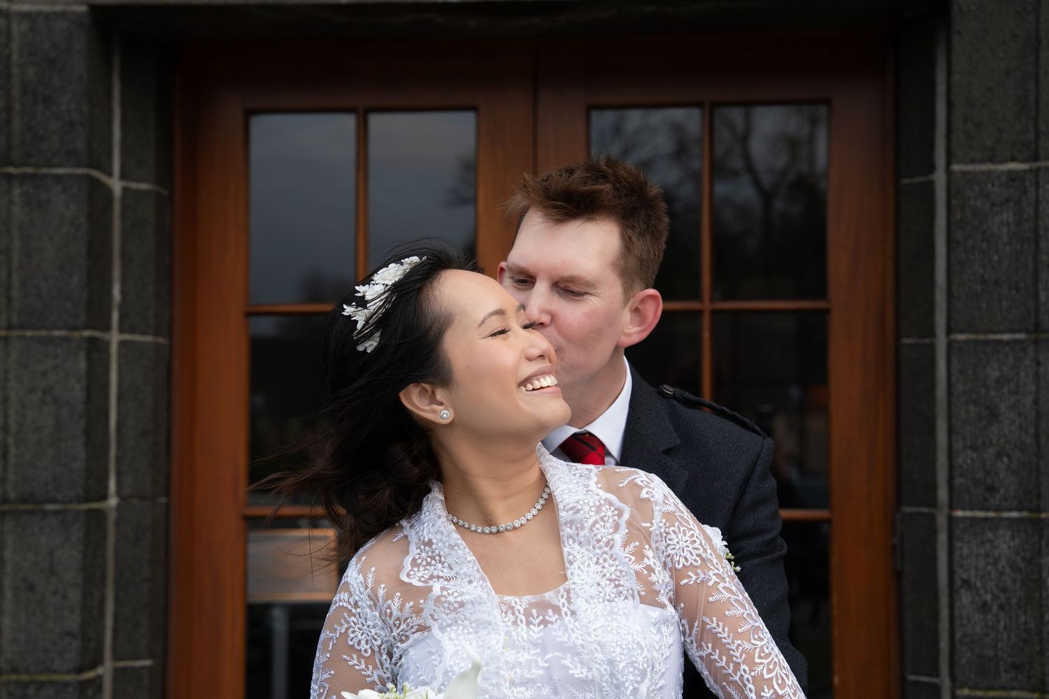 Wedding couple in elegant attire embrace outside historic stone building entrance.