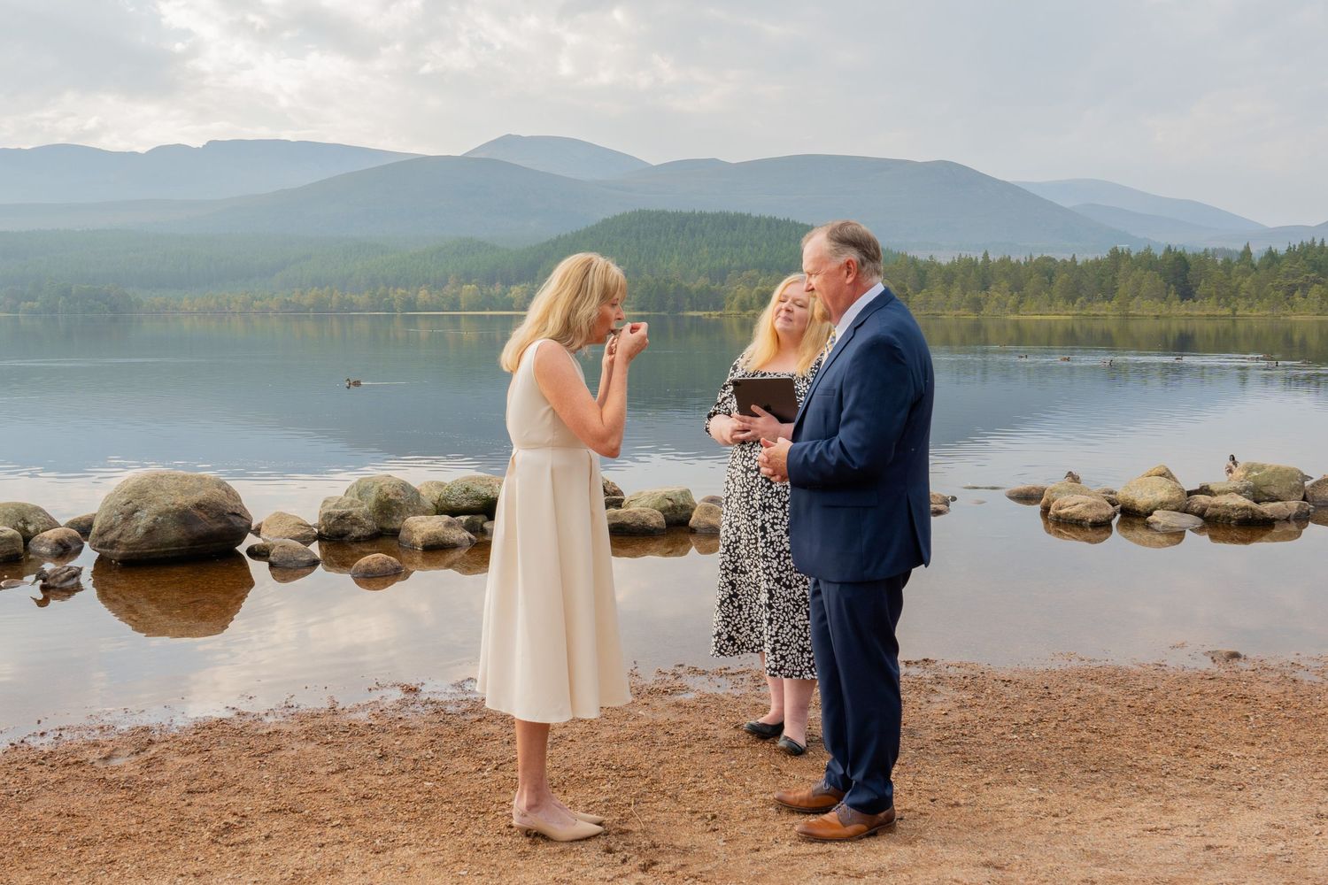 A couple stands together by a serene mountain lake surrounded by rocks and wilderness on their wedding day.