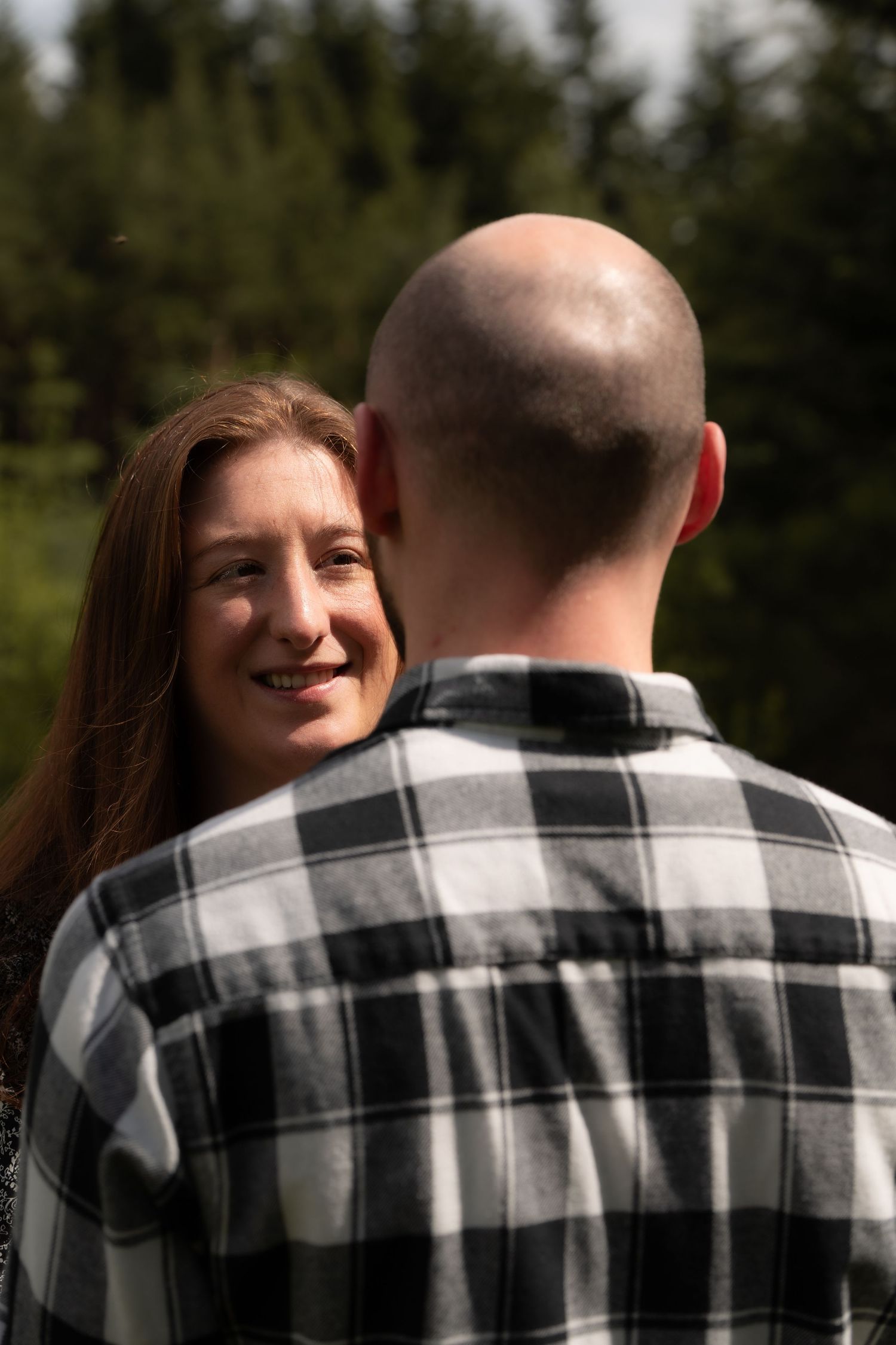 A couple shares an intimate outdoor moment with evergreen trees in the background while one wears a plaid flannel shirt.