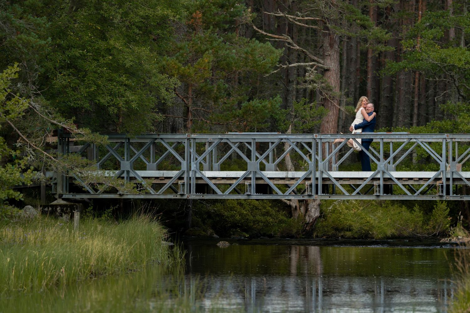 A couple embraces on a metal footbridge over calm water surrounded by green trees and marsh grass.