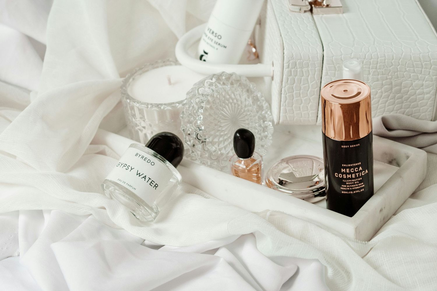 Luxury beauty and skincare products arranged on white fabric with rose gold details and glass bottles.