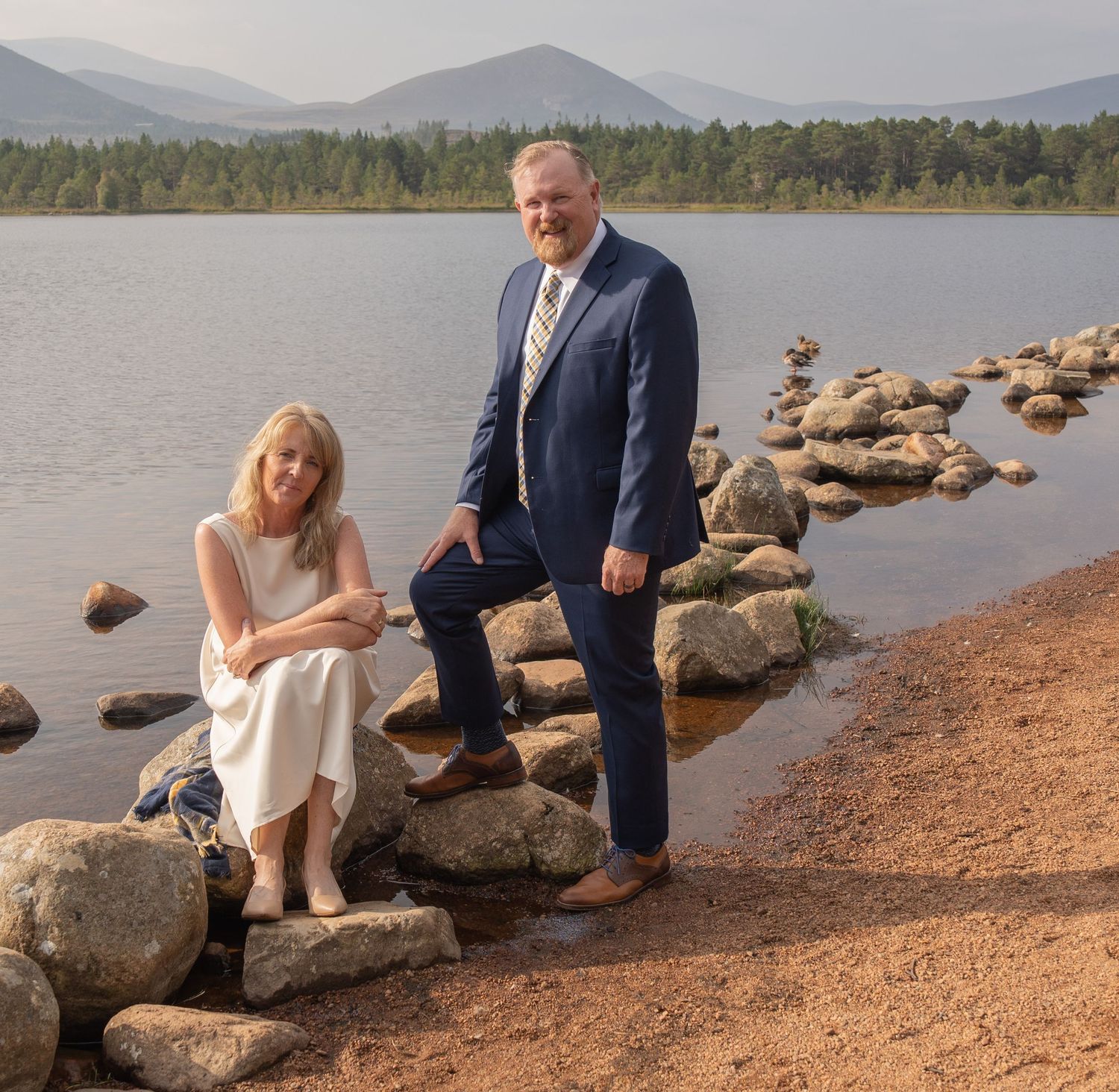 Two people sitting on rocky shoreline near lake with mountains and forest in background.