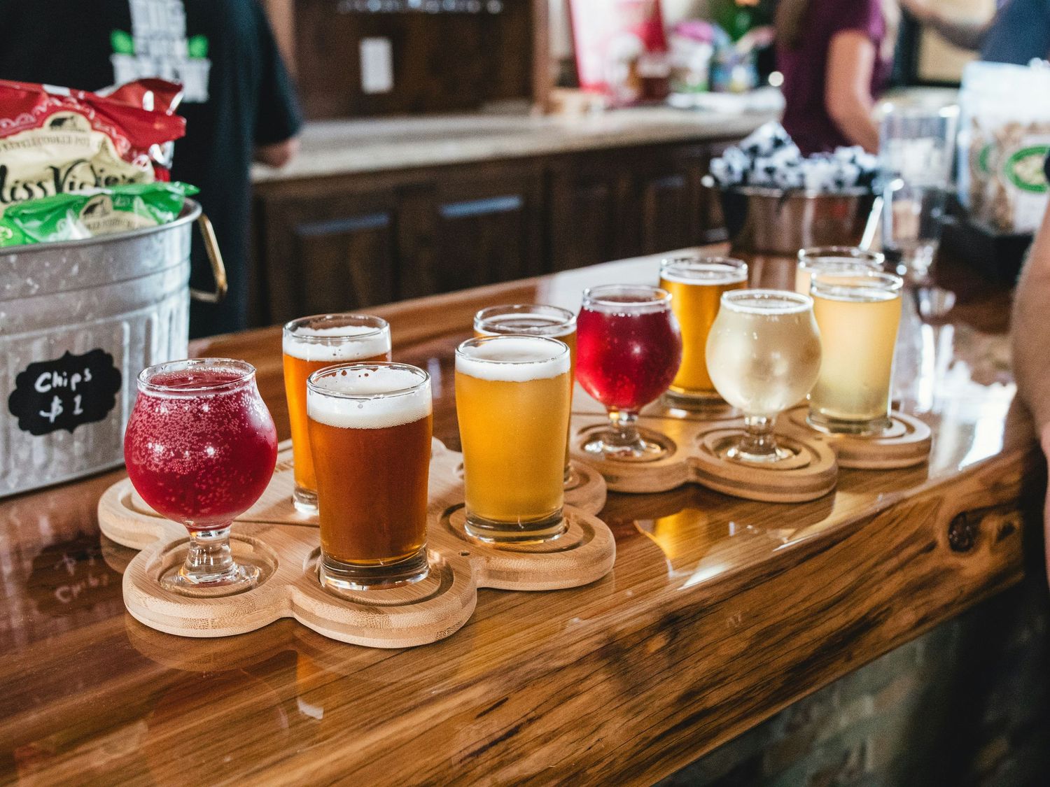 A craft beer flight with six different colored beers ranging from deep red to golden yellow served on wooden coasters.