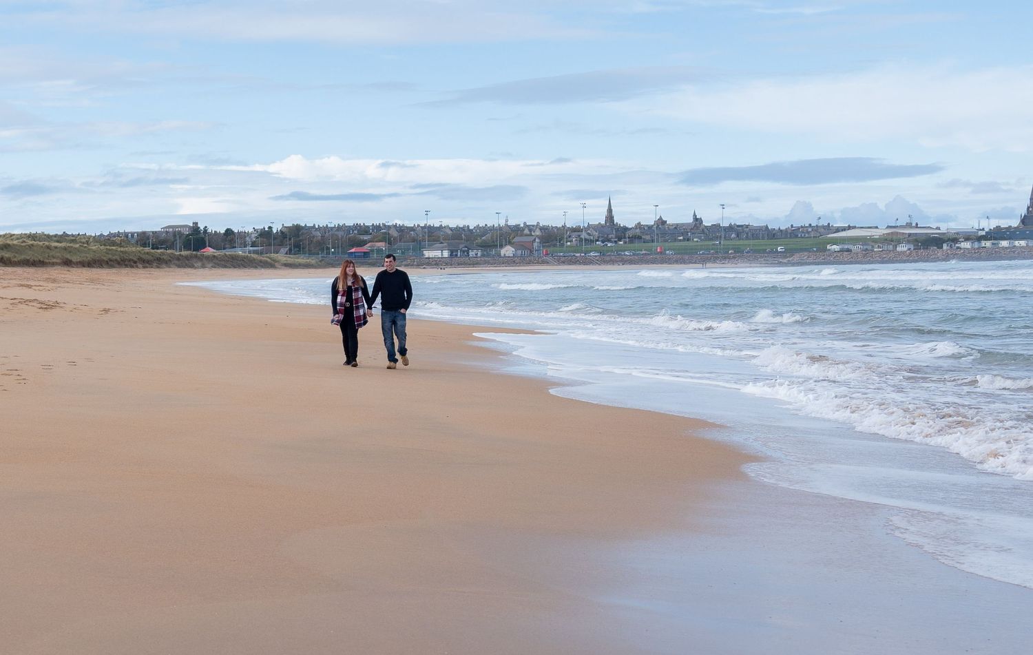 People walk along a scenic coastal beach with rolling waves and dramatic sky in the background.