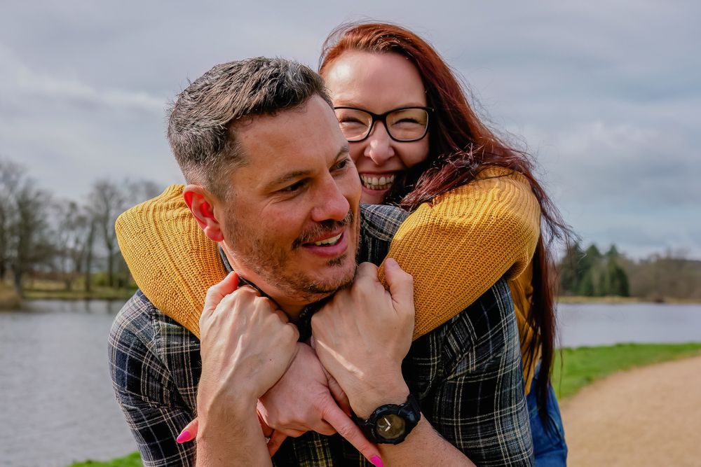 Two people share a joyful embrace and laugh together by a lake on an overcast day.