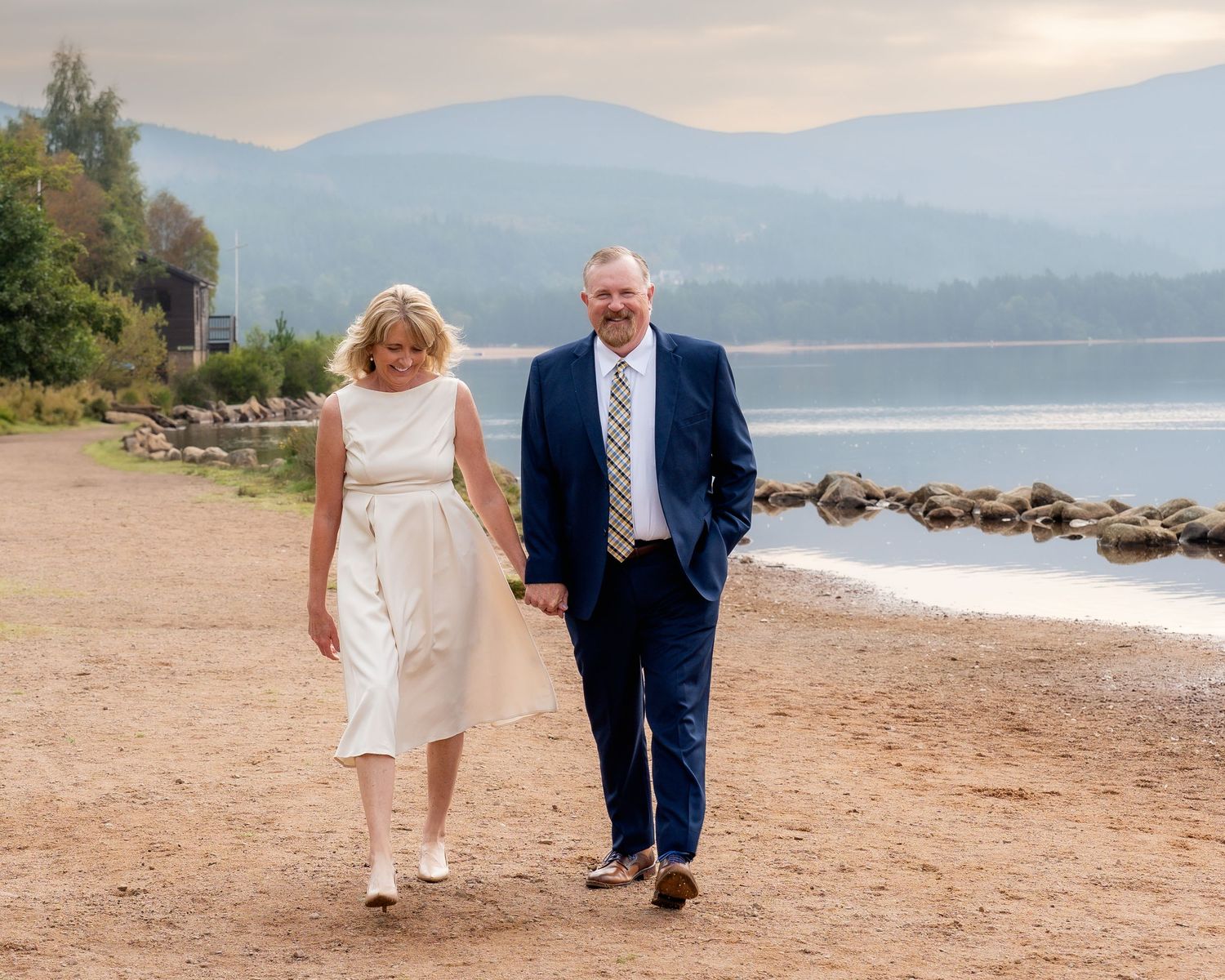 A couple walking together on a waterfront path with mountains and lake in the distance.