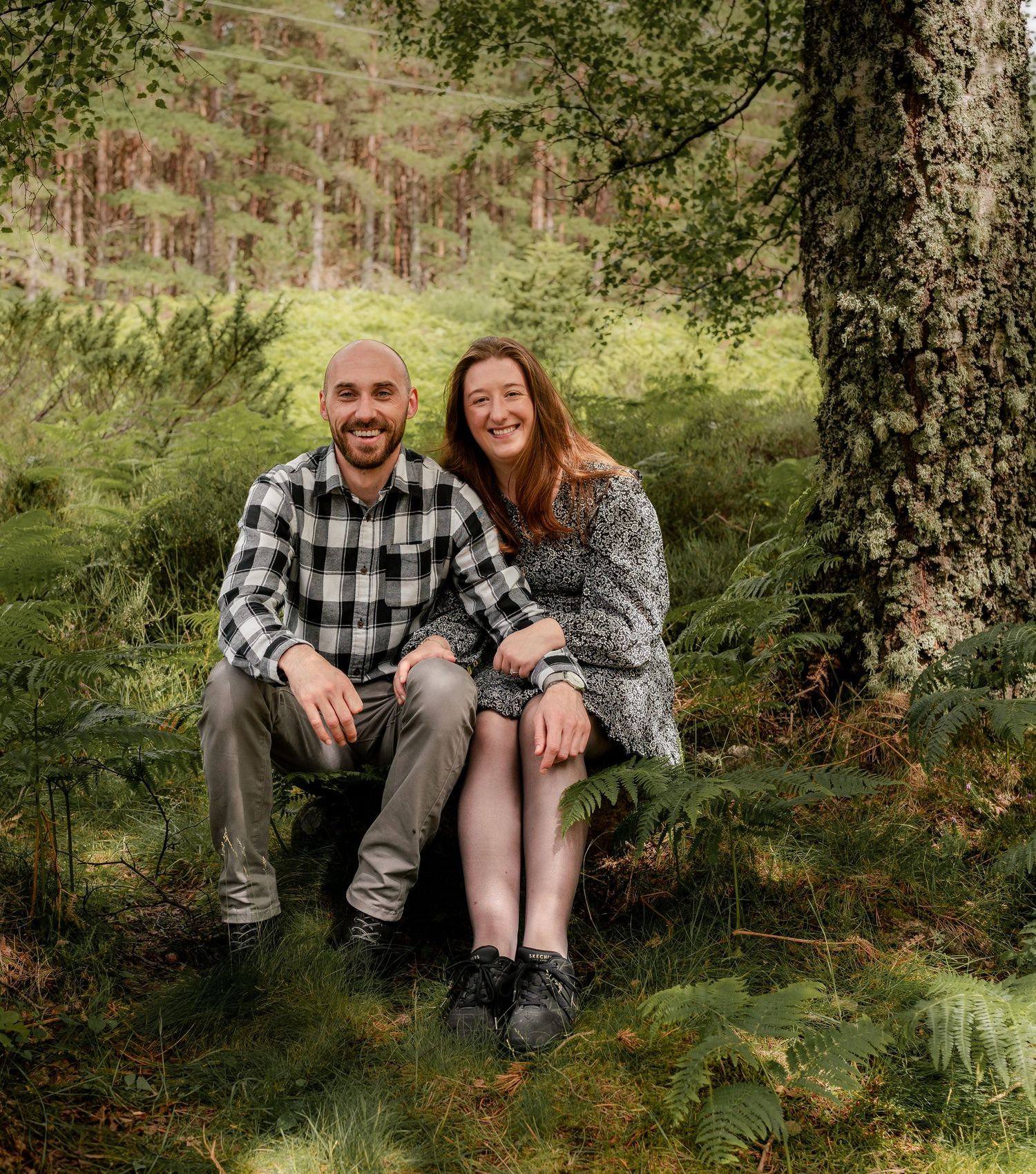 A couple sits together on a mossy log in a lush forest setting with natural greenery surrounding them.