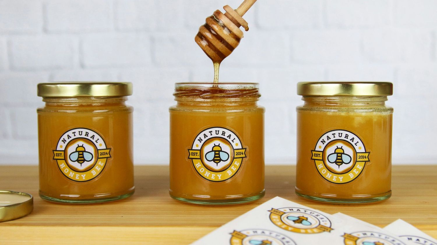 Three glass jars of golden honey with branded labels sit on a wooden surface while honey drips from a wooden dipper above.