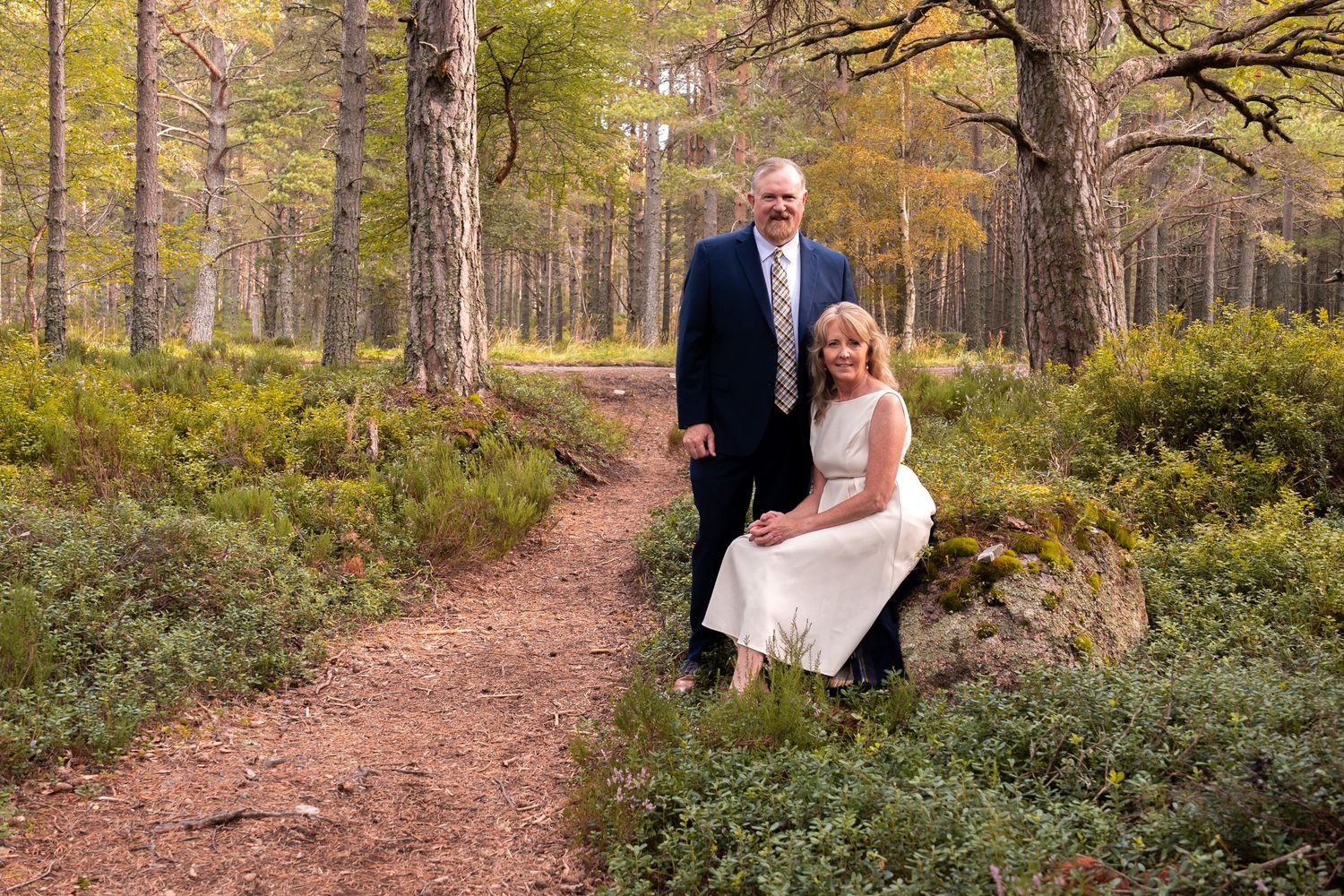 A couple poses together on a wooded forest path during autumn with fall foliage in the background.