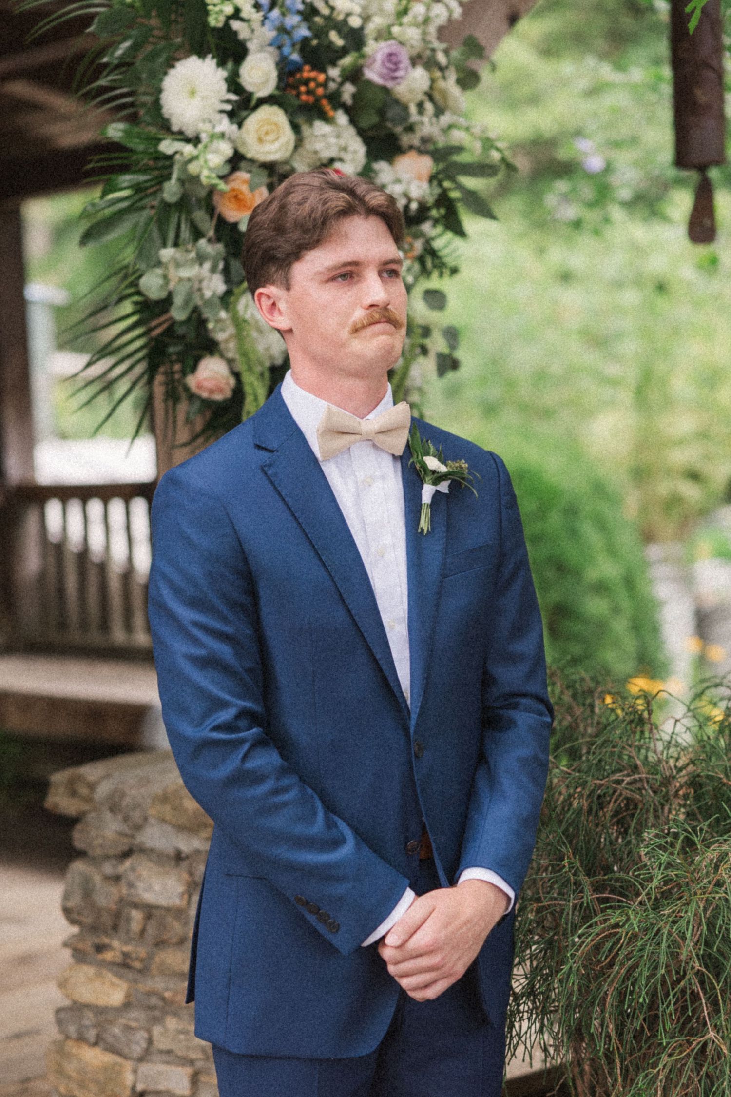 Groom in navy suit and bow tie standing in front of floral wedding arch arrangement.