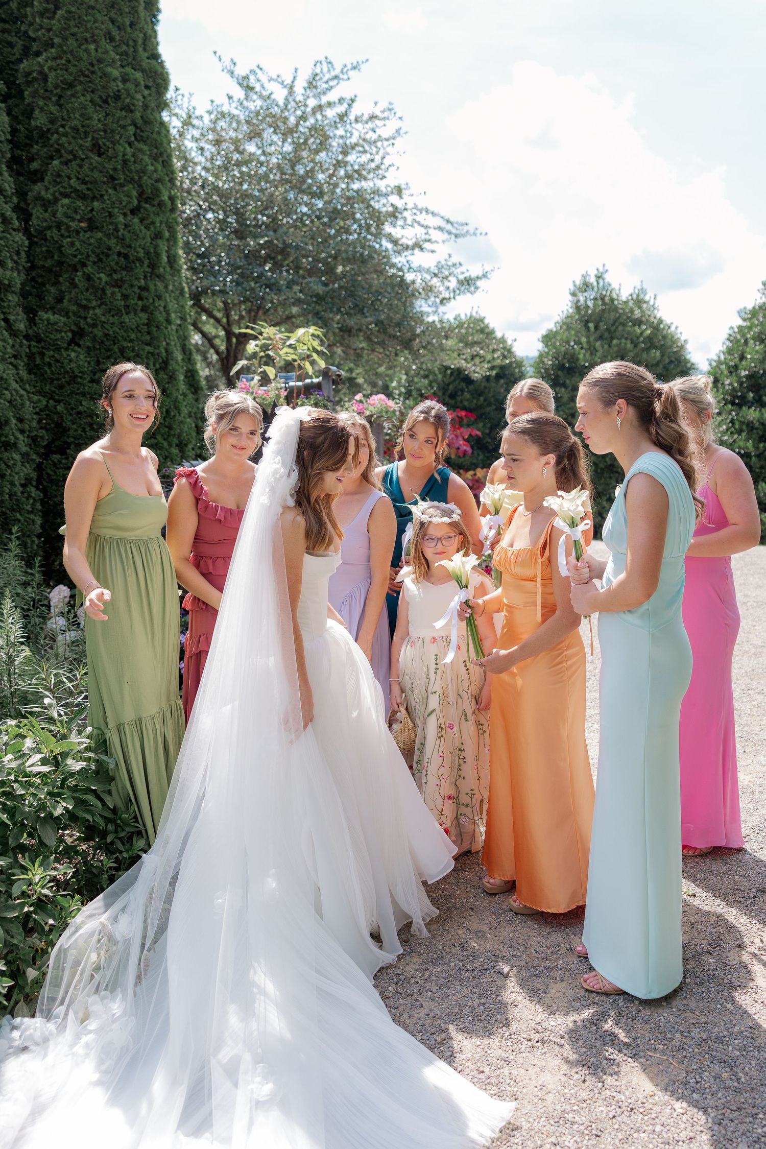 A wedding party in pastel-colored dresses poses together in a garden setting.